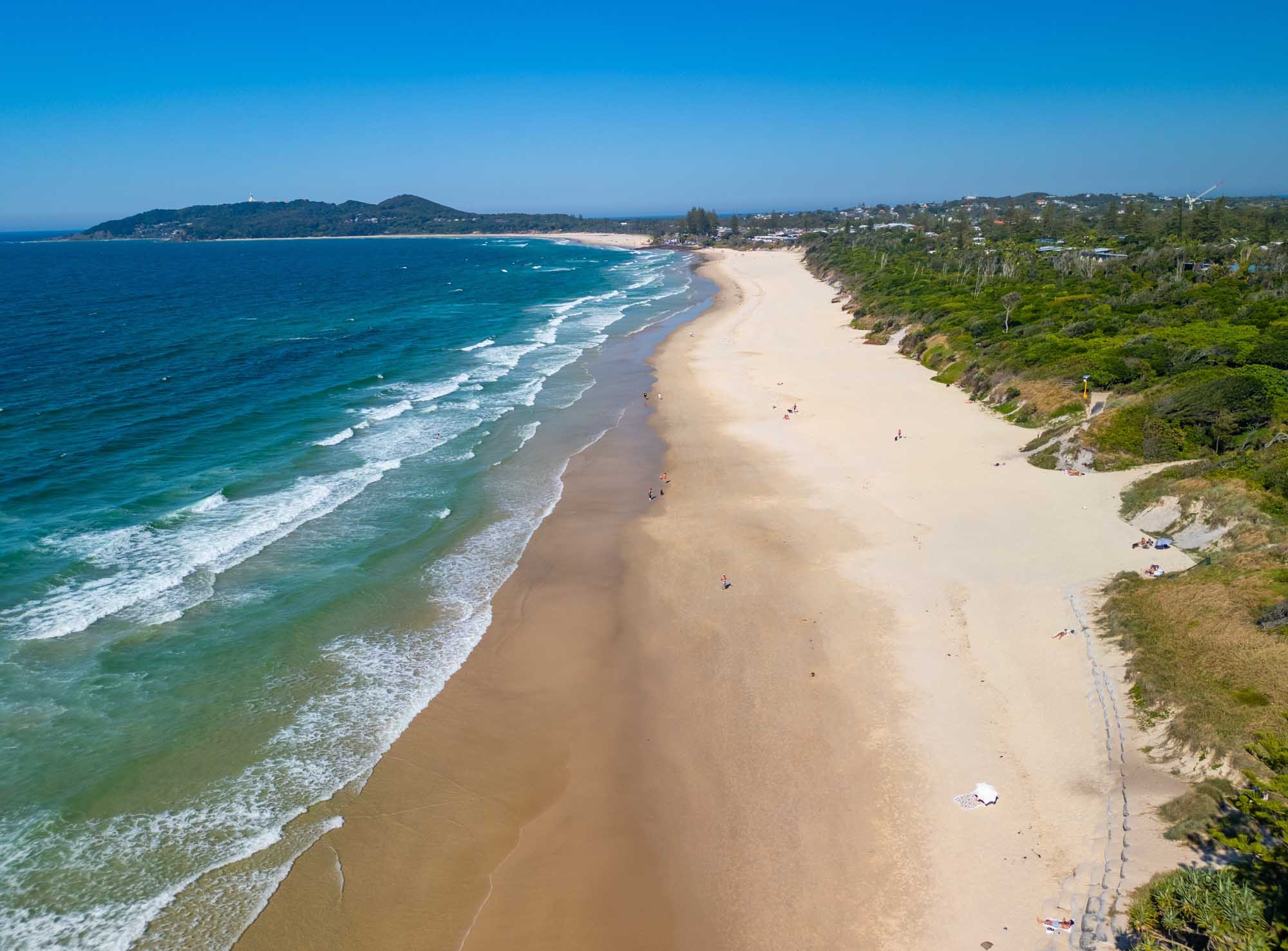 An aerial photograph of Belongil Beach in Byron Bay, New South Wales, showing a pristine sandy coastline with turquoise waters, white-capped waves, and lush green vegetation along the clifftops. The expansive beach features scattered beachgoers and a dog exercise area, with the charming coastal town visible in the background under clear blue skies.