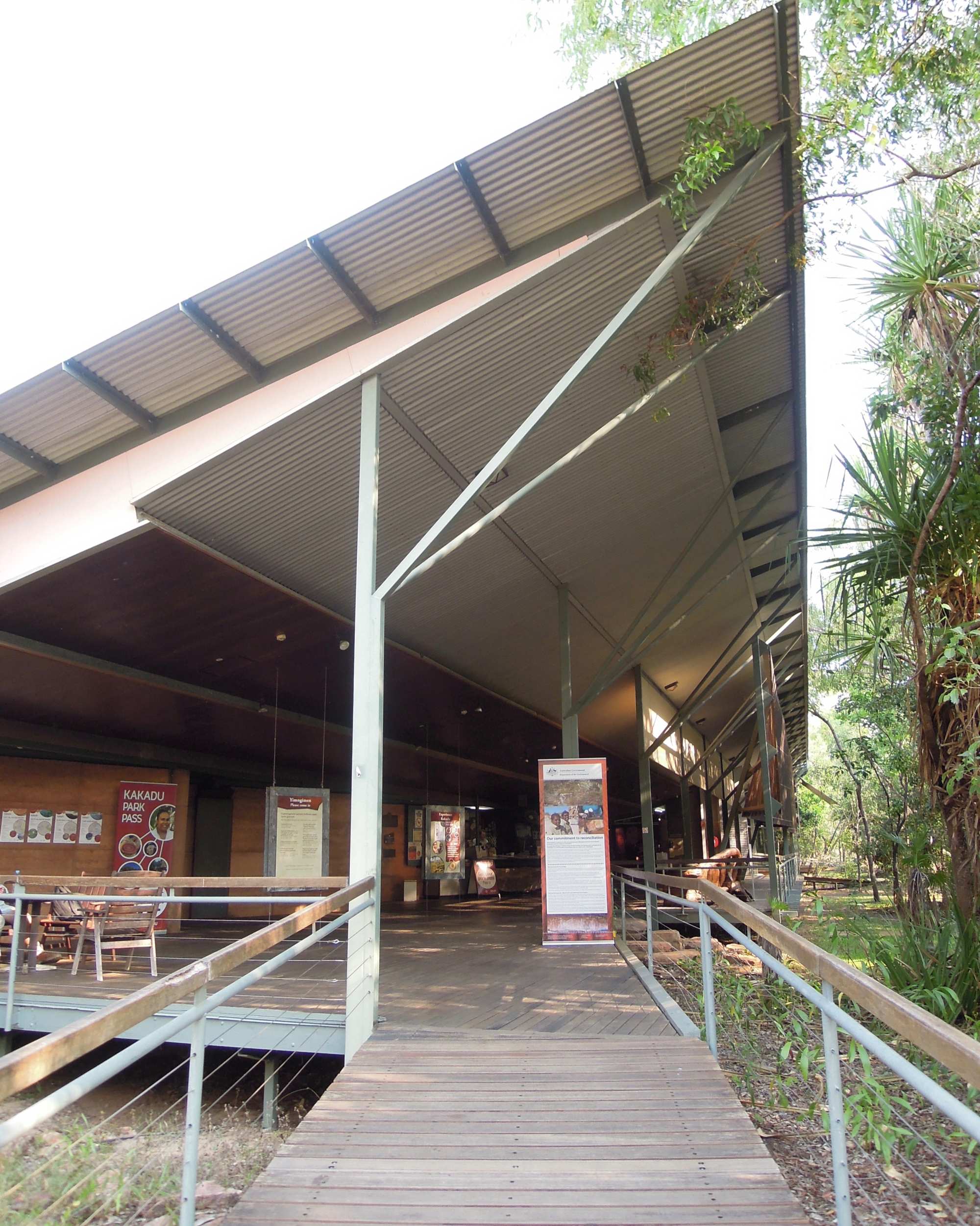 The modern Bowali Visitor Centre at Kakadu National Park features a distinctive architectural design with corrugated metal roofing and wooden boardwalk access. The covered entrance area showcases contemporary sustainable design elements integrated into the natural bushland environment, with native vegetation visible on both sides of the pathway.