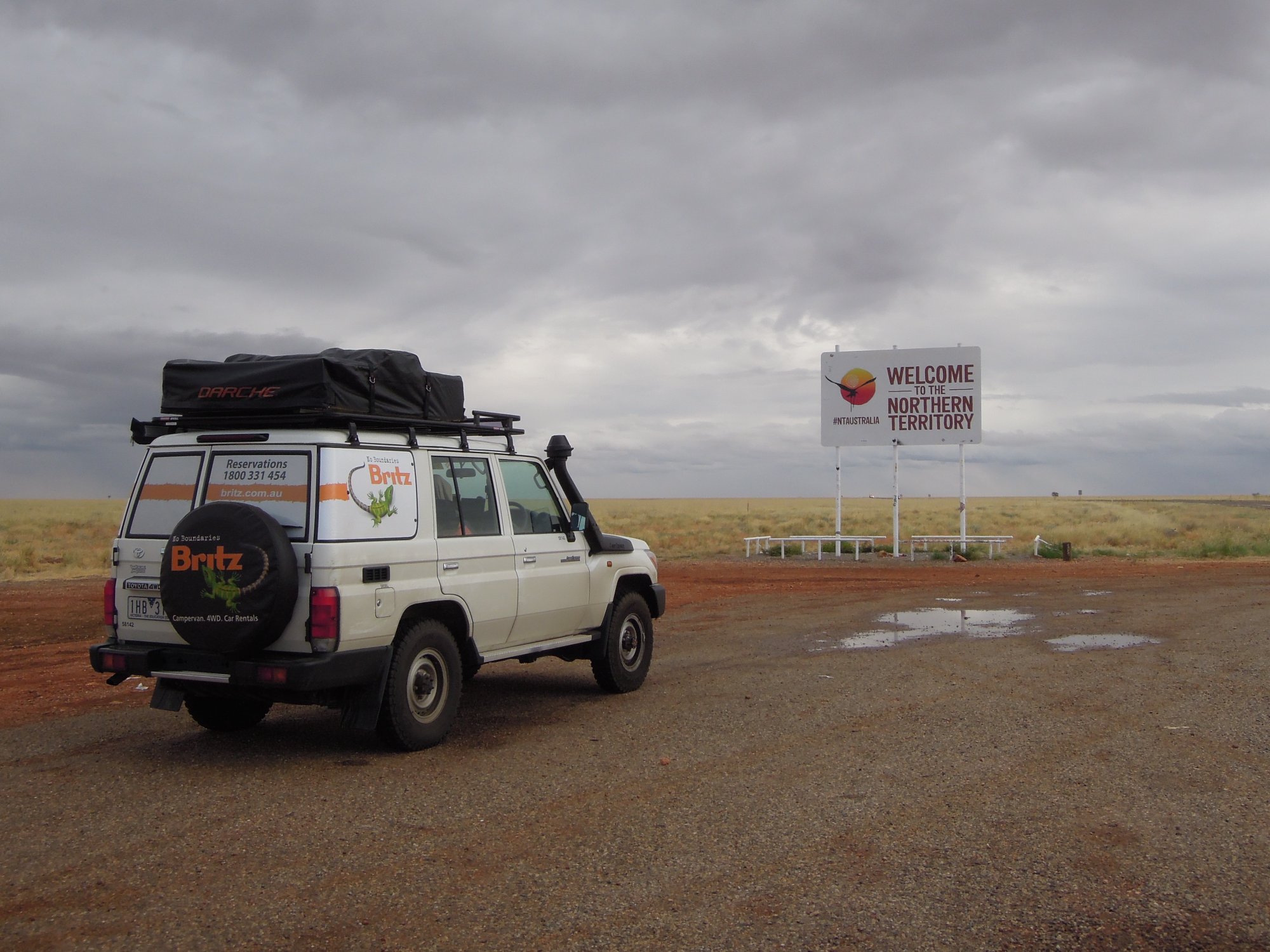 A white Britz rental campervan equipped with a roof rack and black cargo carrier is parked on red earth in front of the official 'Welcome to the Northern Territory' roadside sign, with vast, flat outback landscape and overcast sky in the background.