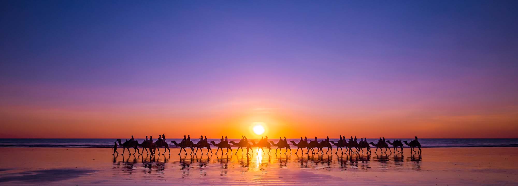 A picturesque sunset scene at Cable Beach in Broome, Western Australia, featuring a caravan of camels and handlers silhouetted against the golden sun reflecting on the wet sand. The dramatic sky transitions from deep blue to warm orange and pink hues, creating a stunning backdrop for this iconic Australian desert coastal experience.