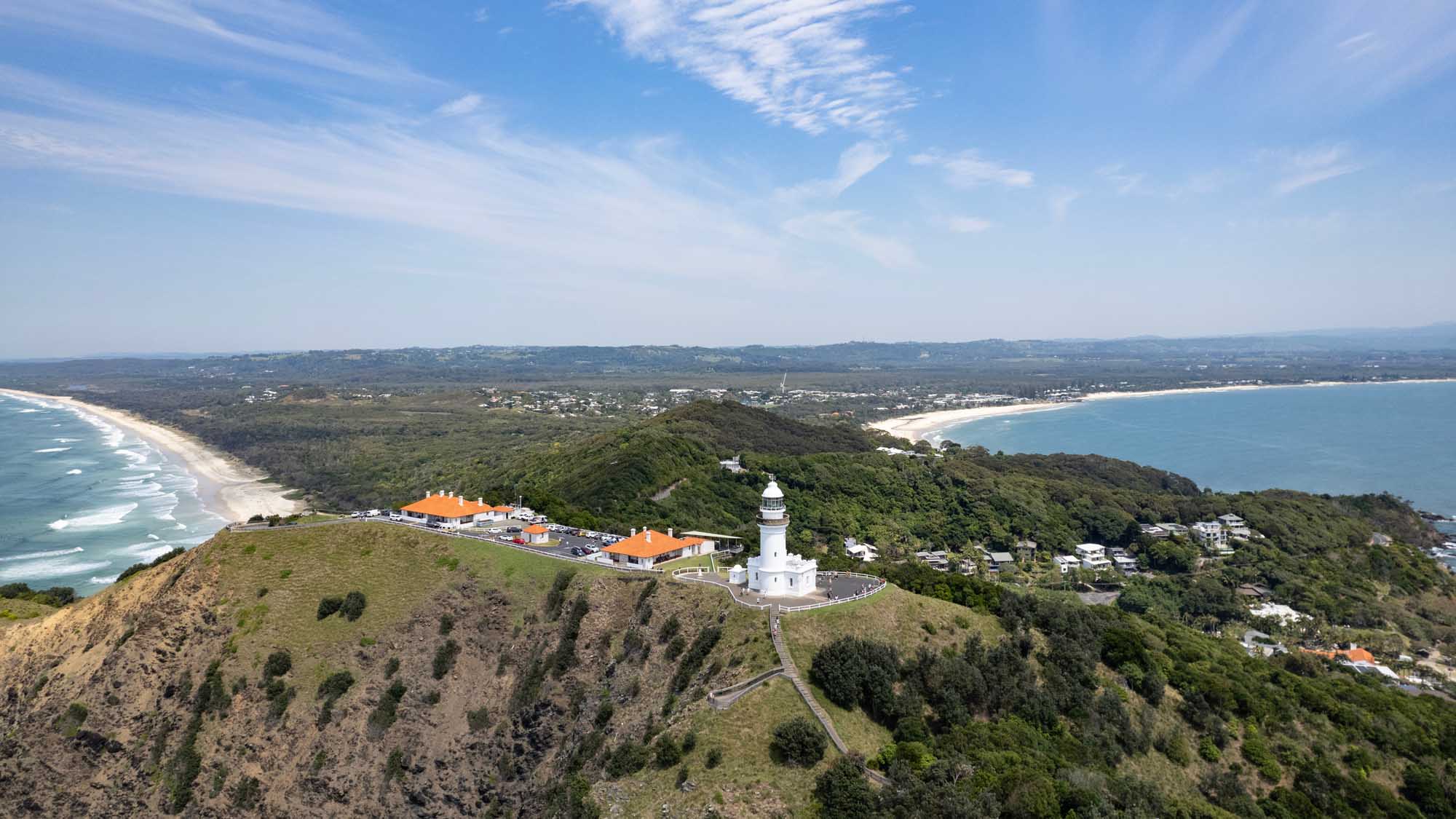 An aerial view of Cape Byron Lighthouse perched on a verdant headland in Byron Bay, New South Wales, with panoramic vistas of pristine beaches, turquoise waters, and the lush hinterland stretching across the horizon. The iconic white lighthouse stands adjacent to orange-roofed visitor facilities, overlooking the curved coastline and residential areas nestled between forested hills and sandy shores.