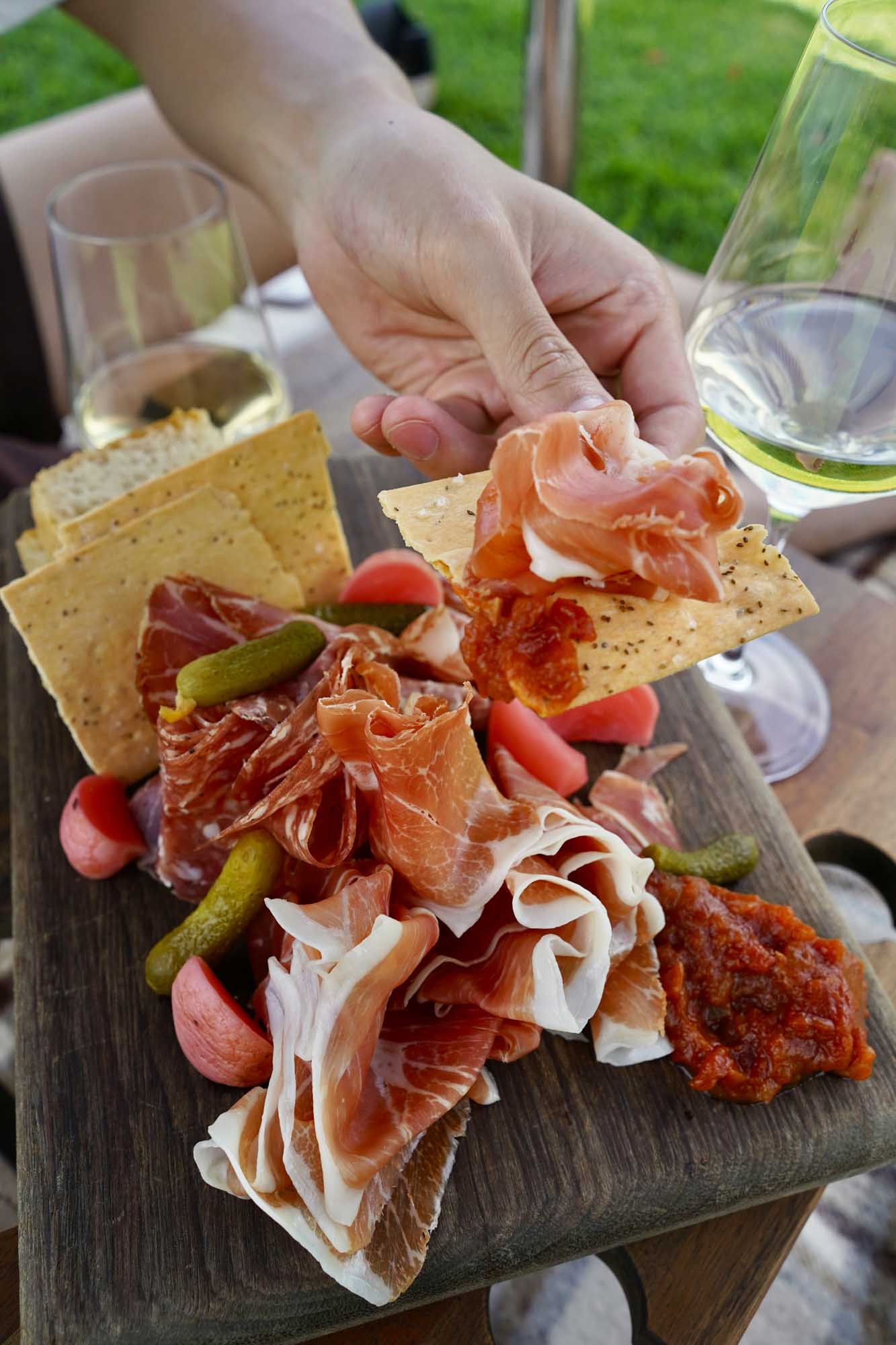 A rustic outdoor charcuterie board featuring an array of sliced cured meats including prosciutto and salami, accompanied by crackers, pickles, cherry tomatoes, and white cheese. The spread is displayed on a dark wooden board with glasses of white wine in the background, set against a lush green garden setting—ideal for an elegant outdoor gathering or wine country experience.