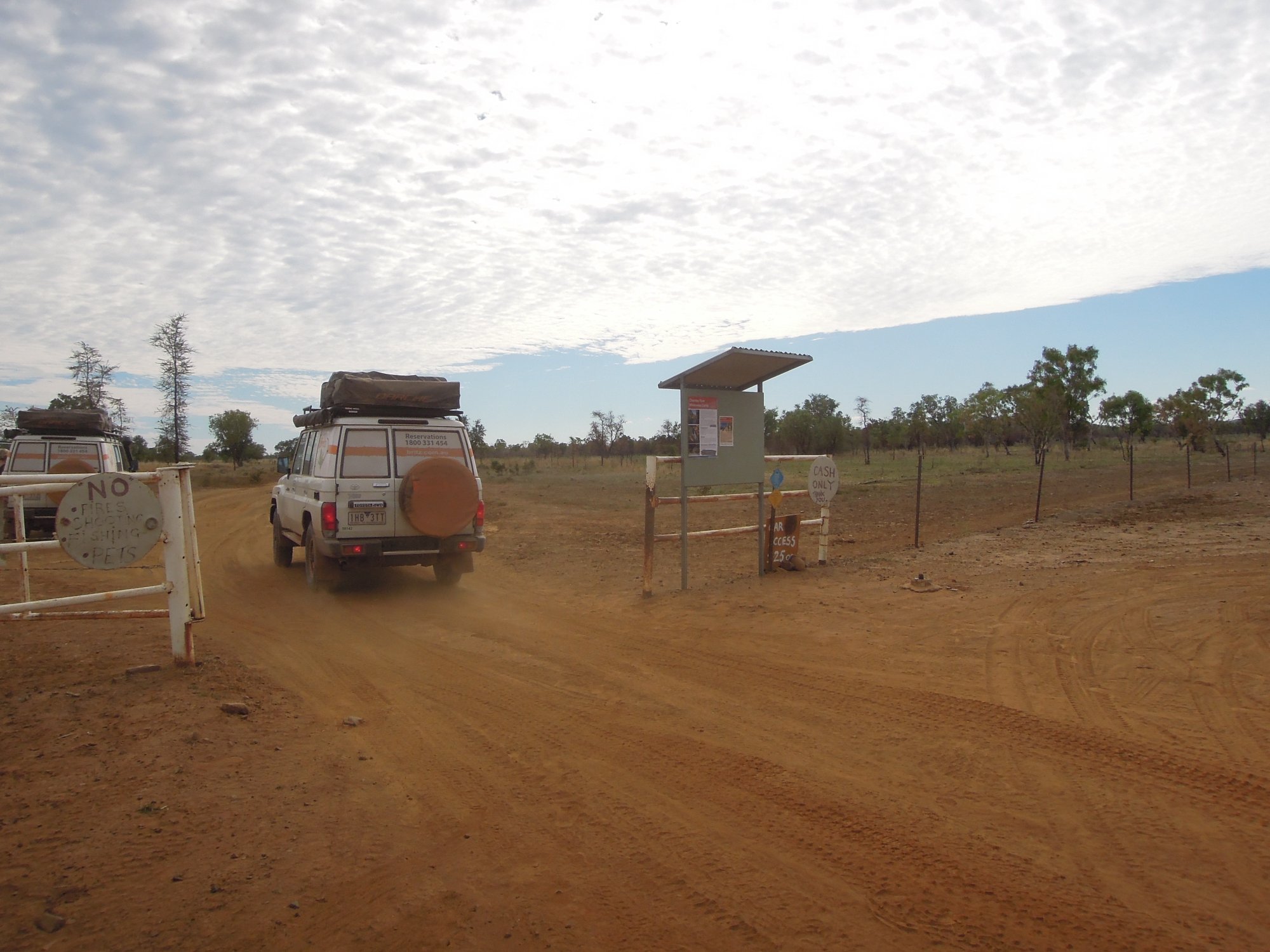 A remote wilderness camp entrance in the Australian Outback, featuring a rustic gate structure with a cash-only payment box, sparse vegetation, and red dirt access road. A white four-wheel-drive vehicle with roof-mounted cargo is visible approaching the gate through the dusty landscape.