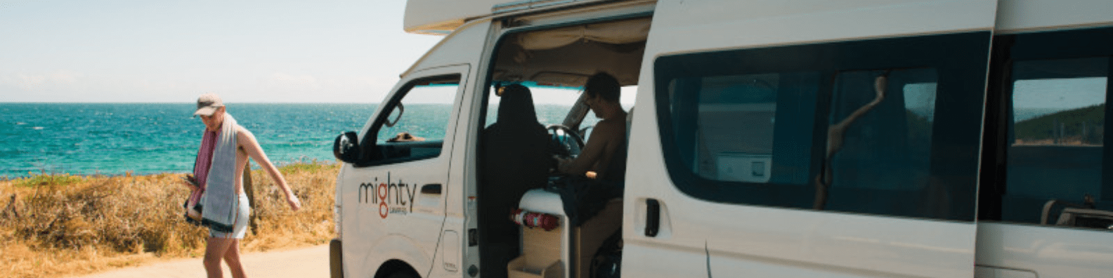 A white campervan branded with 'mighty' is parked on a scenic coastal road overlooking turquoise Mediterranean waters and golden sandy beaches, with passengers boarding for a beach excursion and another visitor walking along the shore.