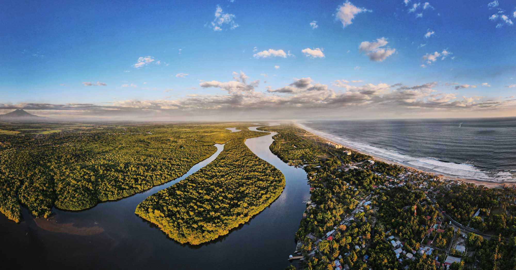 An aerial view of a scenic coastal landscape featuring a meandering river creating distinctive curves through lush green forest, with a developed beachfront community nestled along pristine sandy shores and turquoise ocean waters under a partly cloudy sky.