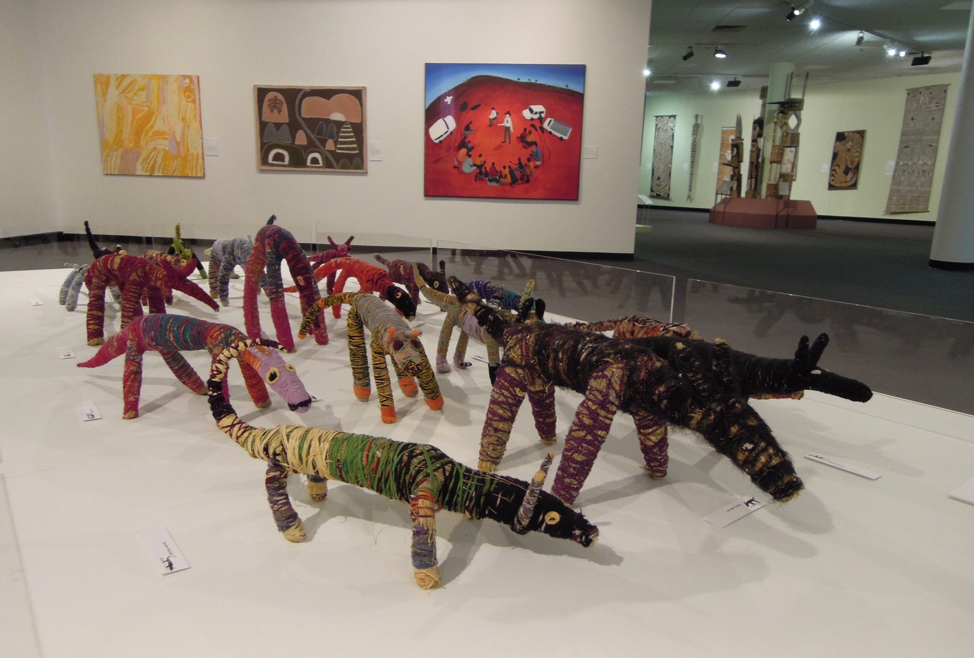 A contemporary art gallery displays a collection of whimsical animal sculptures crafted from vibrant, patterned textiles and fabrics. The figures, which appear to be aardvarks or anteaters, are arranged on a white floor with framed artworks hanging on the walls behind them, creating an engaging and playful exhibition space.