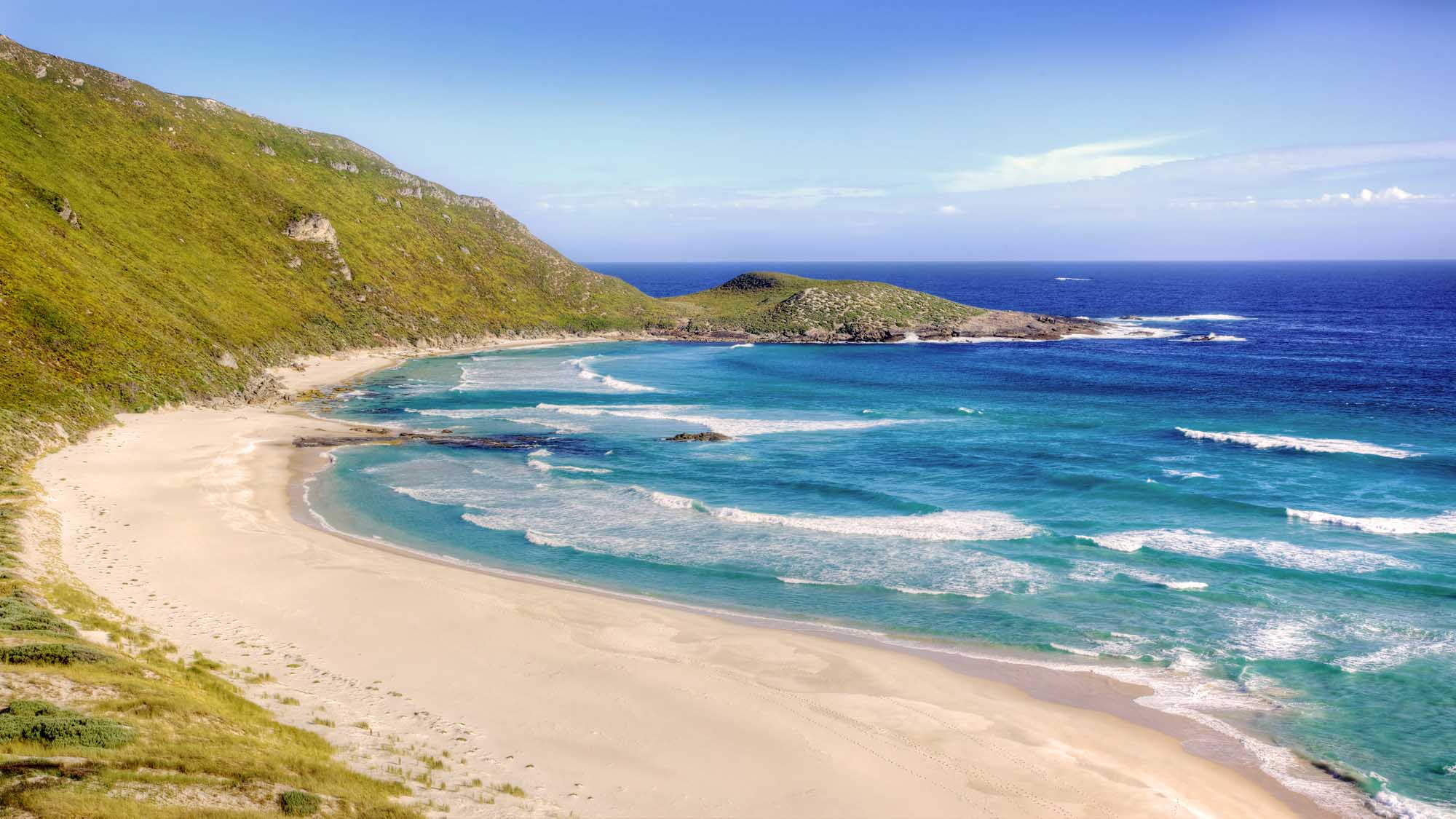 A pristine turquoise cove with white sand beach nestled beneath lush green coastal cliffs in Western Australia. The sheltered bay features gentle waves rolling onto the sandy shore, with rocky outcrops and a small forested headland visible across the water under clear blue skies.