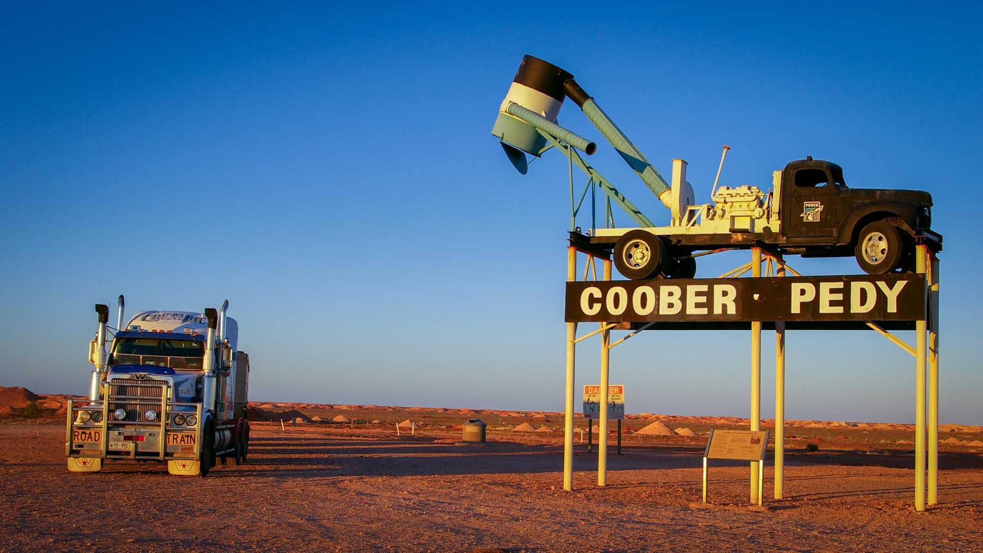 A striking roadside attraction featuring a large mechanical truck with an excavator arm displayed on a yellow steel structure, bearing the Coober Pedy sign. A decorated semi-truck is parked nearby on the red earth landscape under a clear blue sky, exemplifying the quirky desert tourism culture of South Australia's opal mining region.