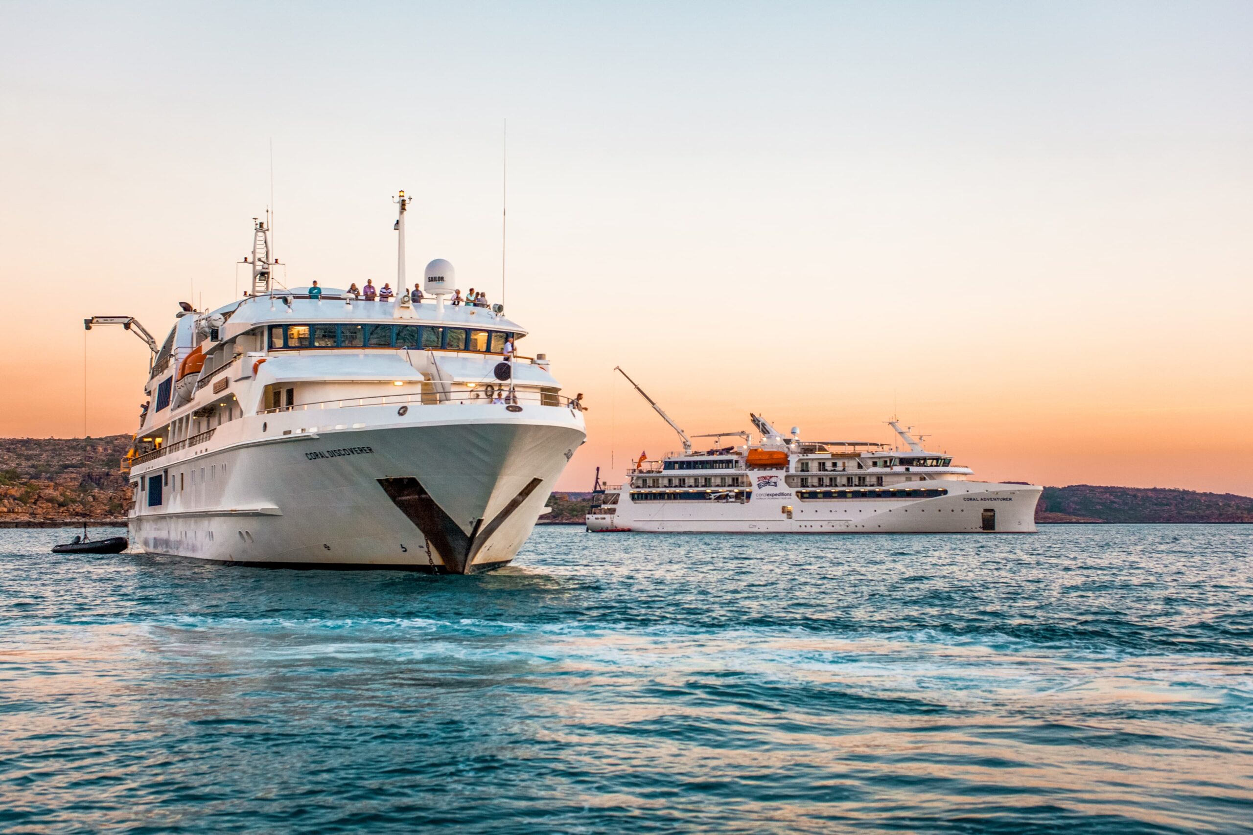 Two expedition cruise vessels, the Coral Adventurer and Coral Discoverer, are anchored together in the turquoise waters of Australia's Kimberley region during a 2019 voyage. The larger ship in the foreground displays passengers on its upper decks, while the second vessel is positioned nearby, with a scenic rocky coastline and golden sunset sky in the background.