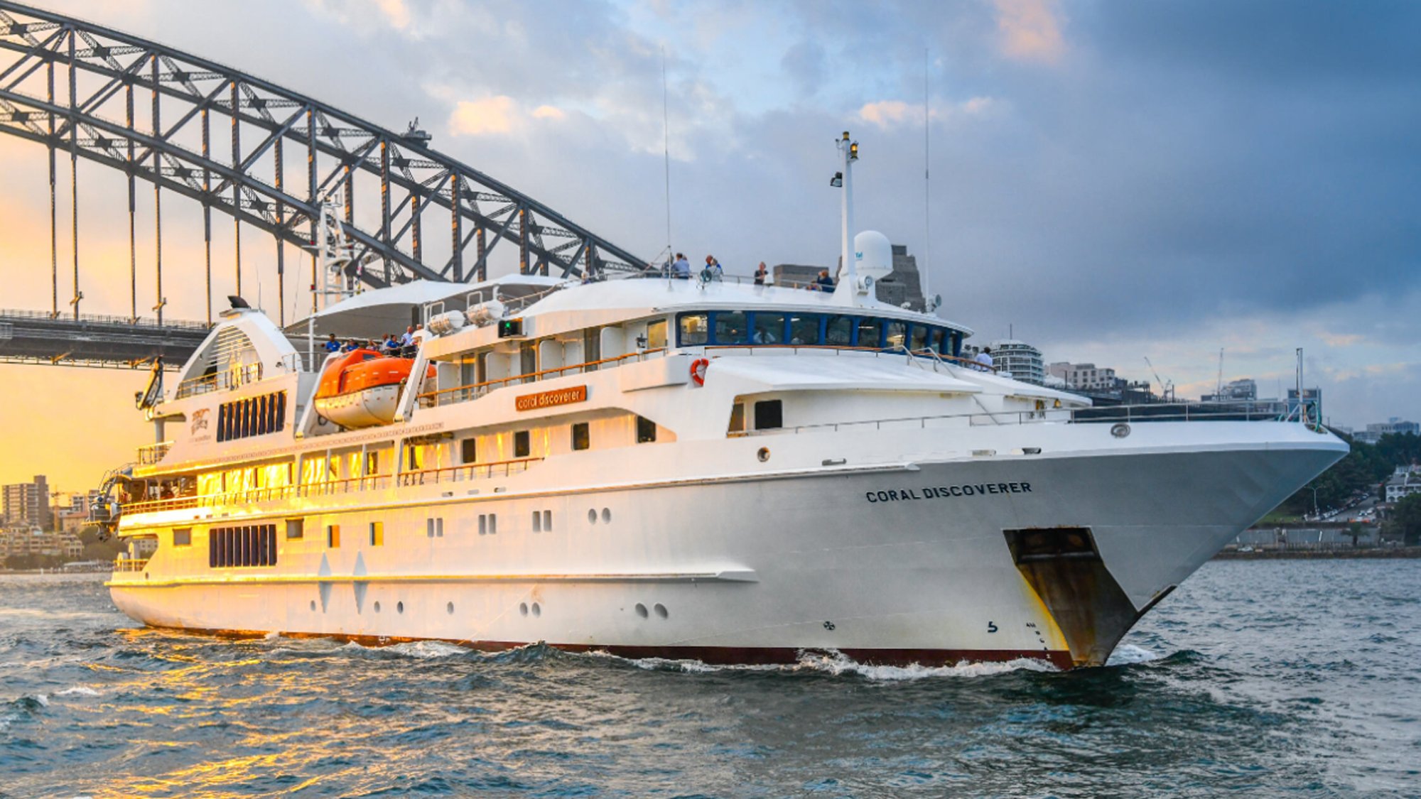 The Coral Discoverer, an expedition cruise vessel, is photographed during golden hour near a large bridge structure, likely in Sydney Harbour. The ship features distinctive yellow and white hull colors with multiple decks and modern amenities typical of small luxury cruise ships.