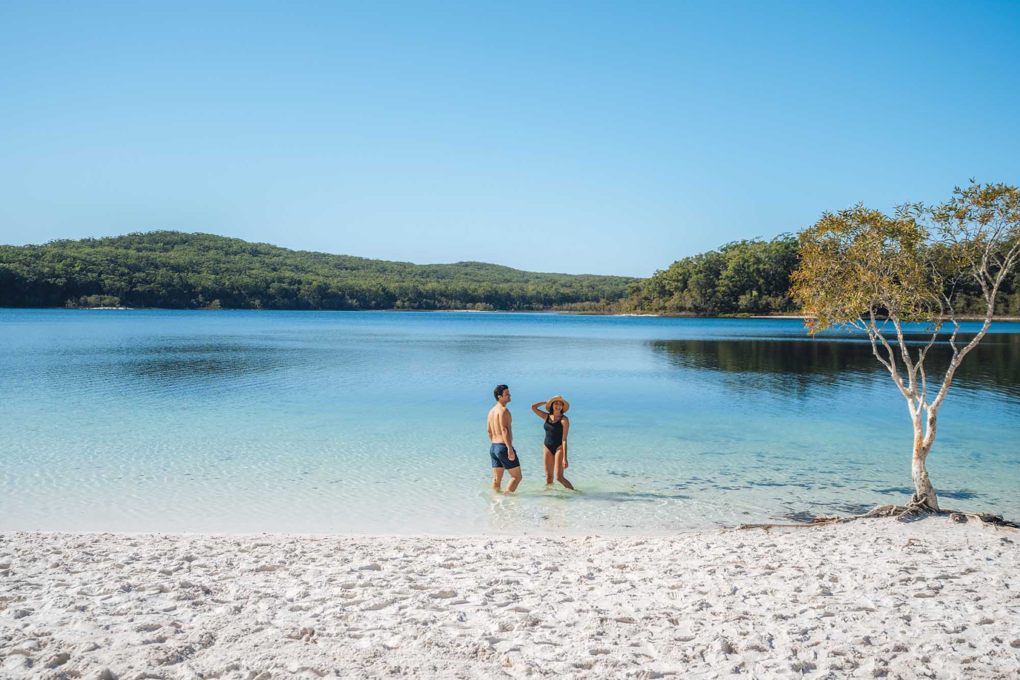 A couple wades in the crystal-clear turquoise waters of Lake McKenzie, a pristine freshwater lake surrounded by lush eucalyptus forests and white sandy beaches. The serene scene captures the natural beauty of this popular destination on Fraser Island, Queensland.