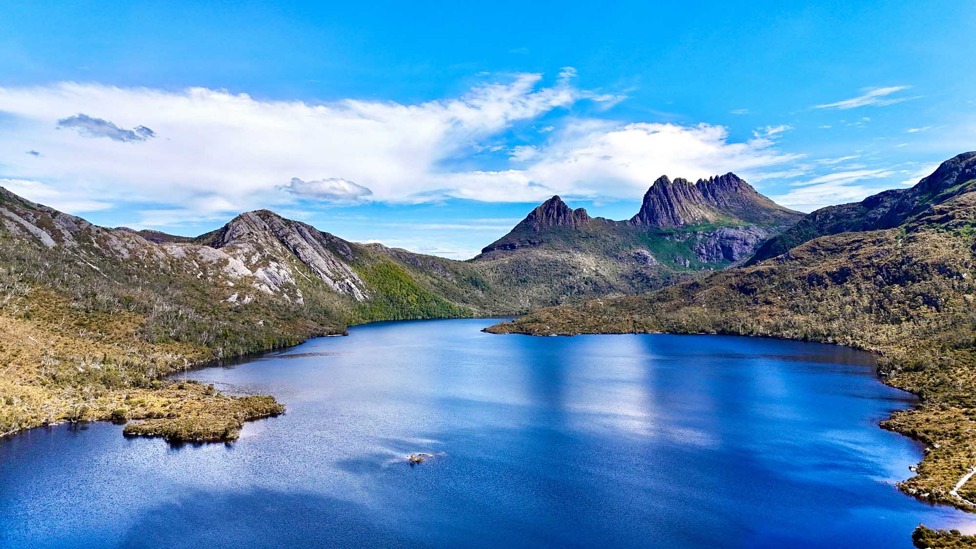 A stunning alpine landscape featuring the distinctive jagged peaks of Cradle Mountain rising majestically above the crystal-clear waters of Dove Lake, surrounded by native forest and moorland vegetation under a bright blue sky with scattered clouds.