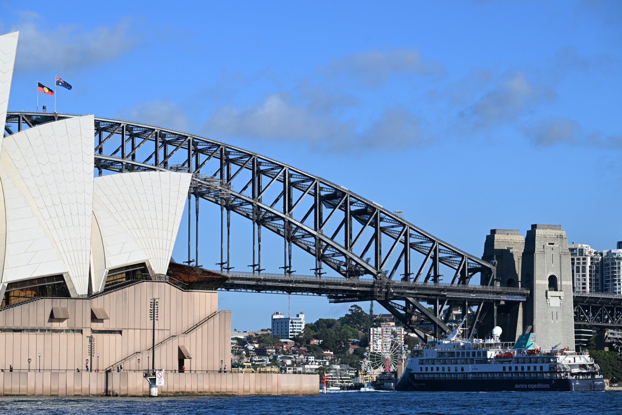 The expedition cruise ship Douglas Mawson dwarfed by the iconic Sydney Harbour Bridge and Sydney Opera House. The blue-hulled vessel is moored in the iconic natural harbour with the bridge's distinctive steel arch structure and the Opera House's white shell-shaped roof prominently featured against a clear blue sky.