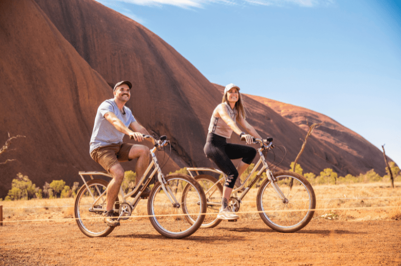 Two cyclists ride bikes on red desert terrain with the distinctive massive sandstone monolith of Uluru rising dramatically in the background under clear blue skies. This iconic Australian landmark and UNESCO World Heritage Site is set in the arid Red Centre landscape.
