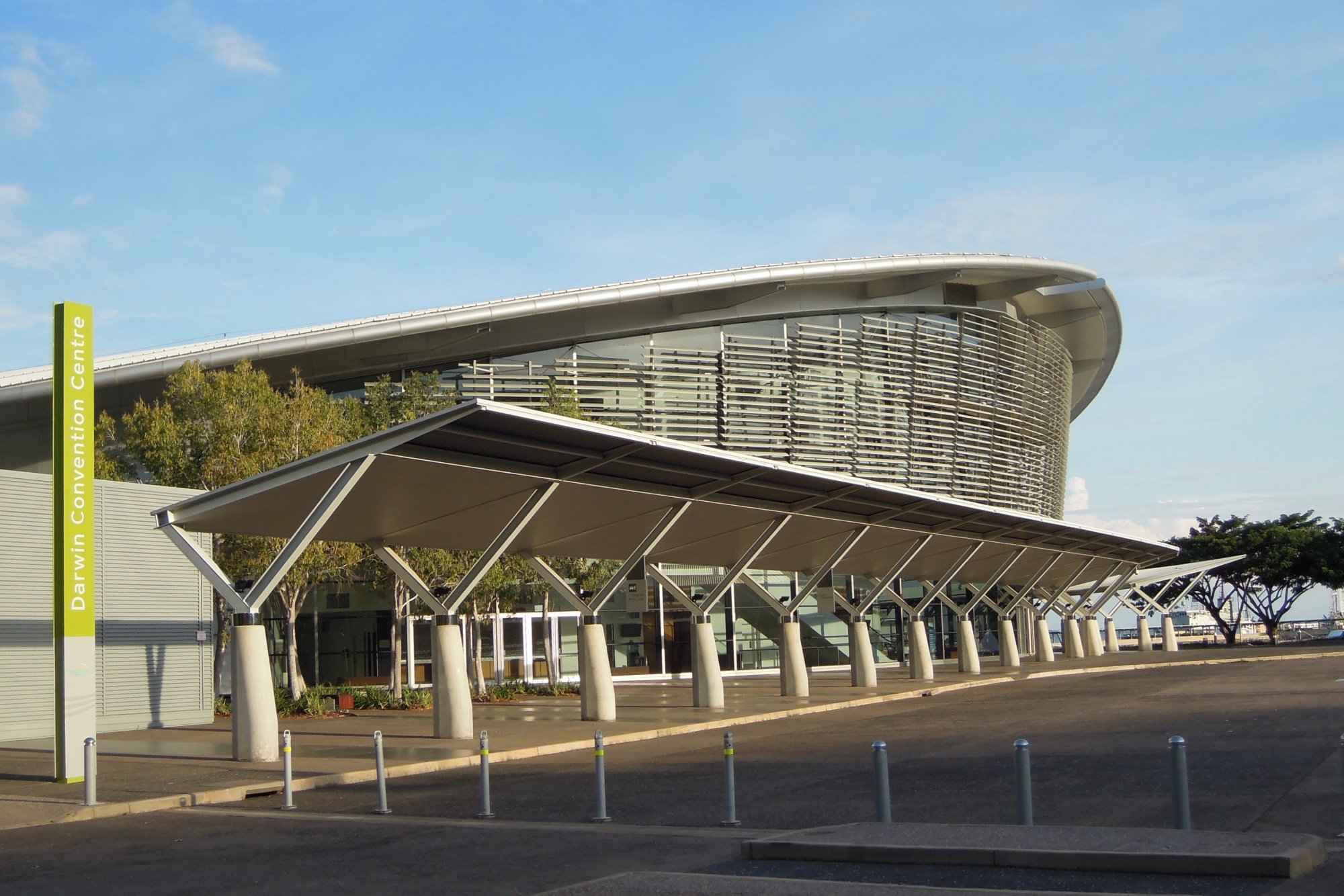 The Darwin Convention Centre features striking contemporary architecture with a distinctive curved metallic roof, angled support columns, and louvered sun-screening elements. The building's sleek design showcases modern civic infrastructure in Australia's Northern Territory.