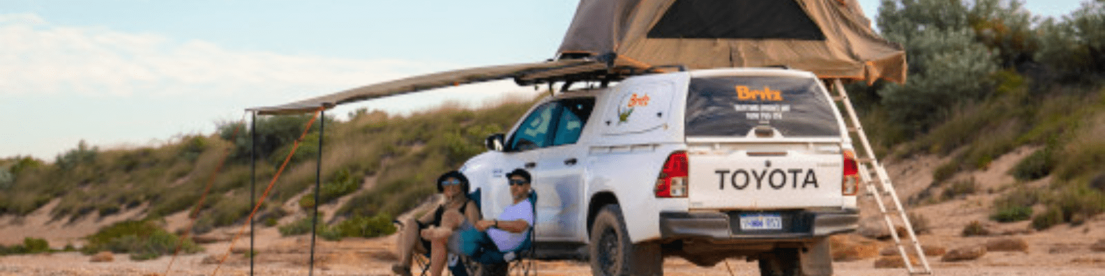 A white Toyota pickup truck equipped with a tan canvas rooftop tent serves as a mobile camping base in an arid desert landscape. Two people relax in camping chairs beneath the extended awning, enjoying the outdoor camping experience in what appears to be a remote desert region with sparse vegetation and rocky terrain.