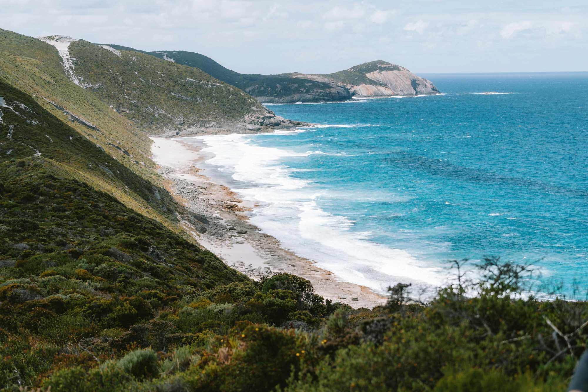 A stunning aerial view of rugged green coastal cliffs meeting a pristine white sandy beach and turquoise waters near Albany, Western Australia. The landscape features steep vegetated hillsides, a secluded cove with breaking waves, and rocky headlands extending into the Indian Ocean under a partly cloudy sky.