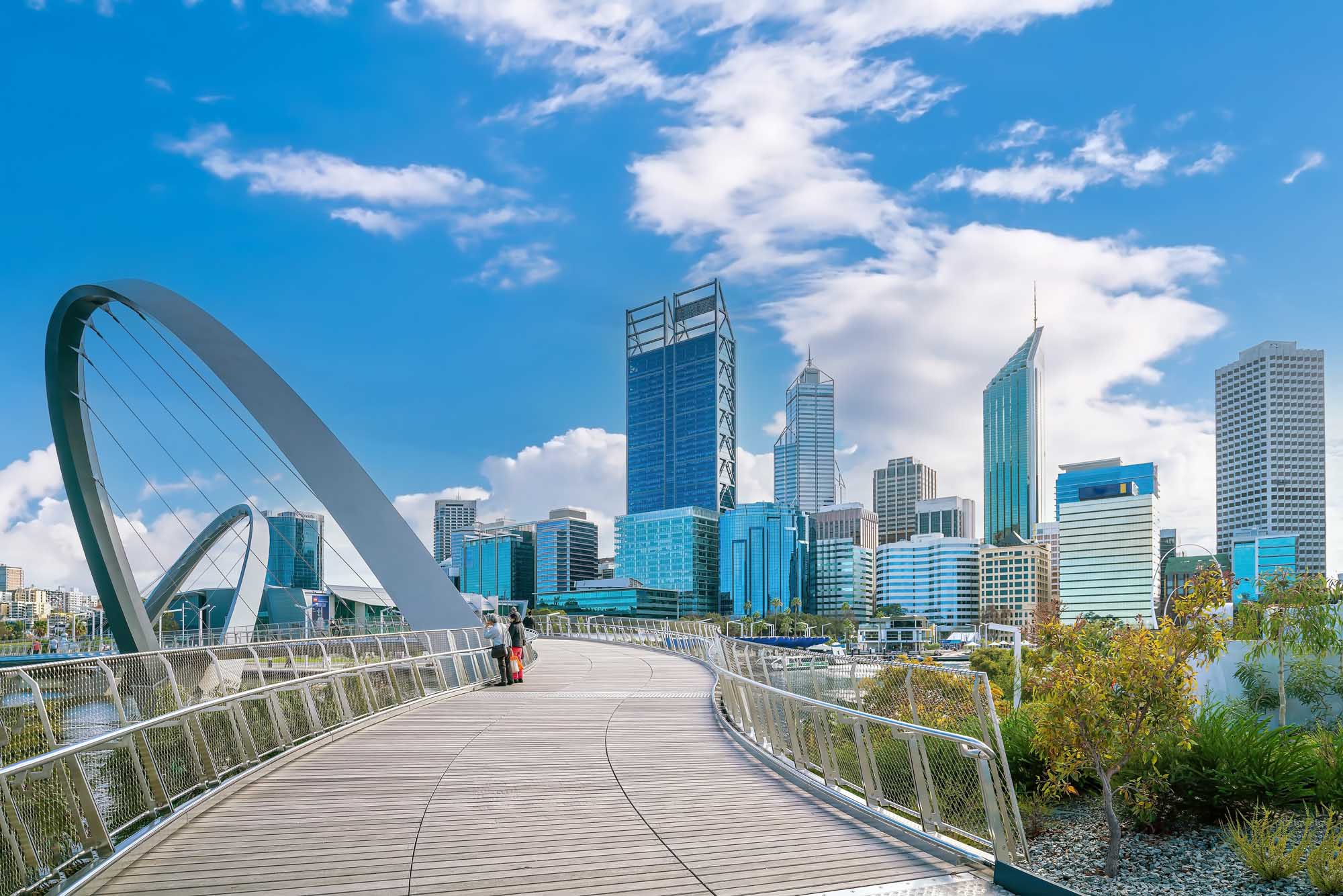 A striking view of Perth's modern downtown skyline framed by the distinctive curved architecture of the Elizabeth Quay Bridge, featuring blue glass towers and contemporary high-rises against a bright blue sky with white clouds. The pedestrian bridge offers an elevated vantage point over the Swan River waterfront.