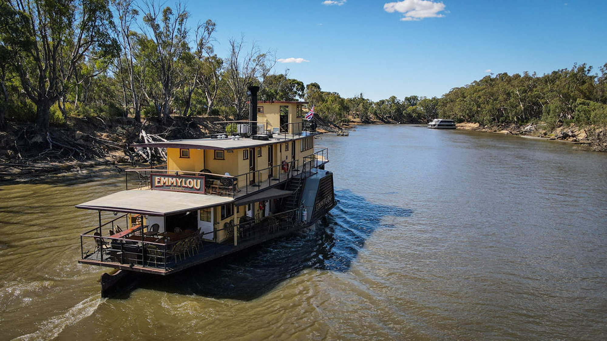 A serene riverside scene at golden hour showing the Murray River with lush green vegetation lining both banks, viewed from a covered deck or balcony of what appears to be a river cruise vessel. The golden sunlight reflects across the calm water, creating a peaceful atmosphere ideal for river cruise travel.