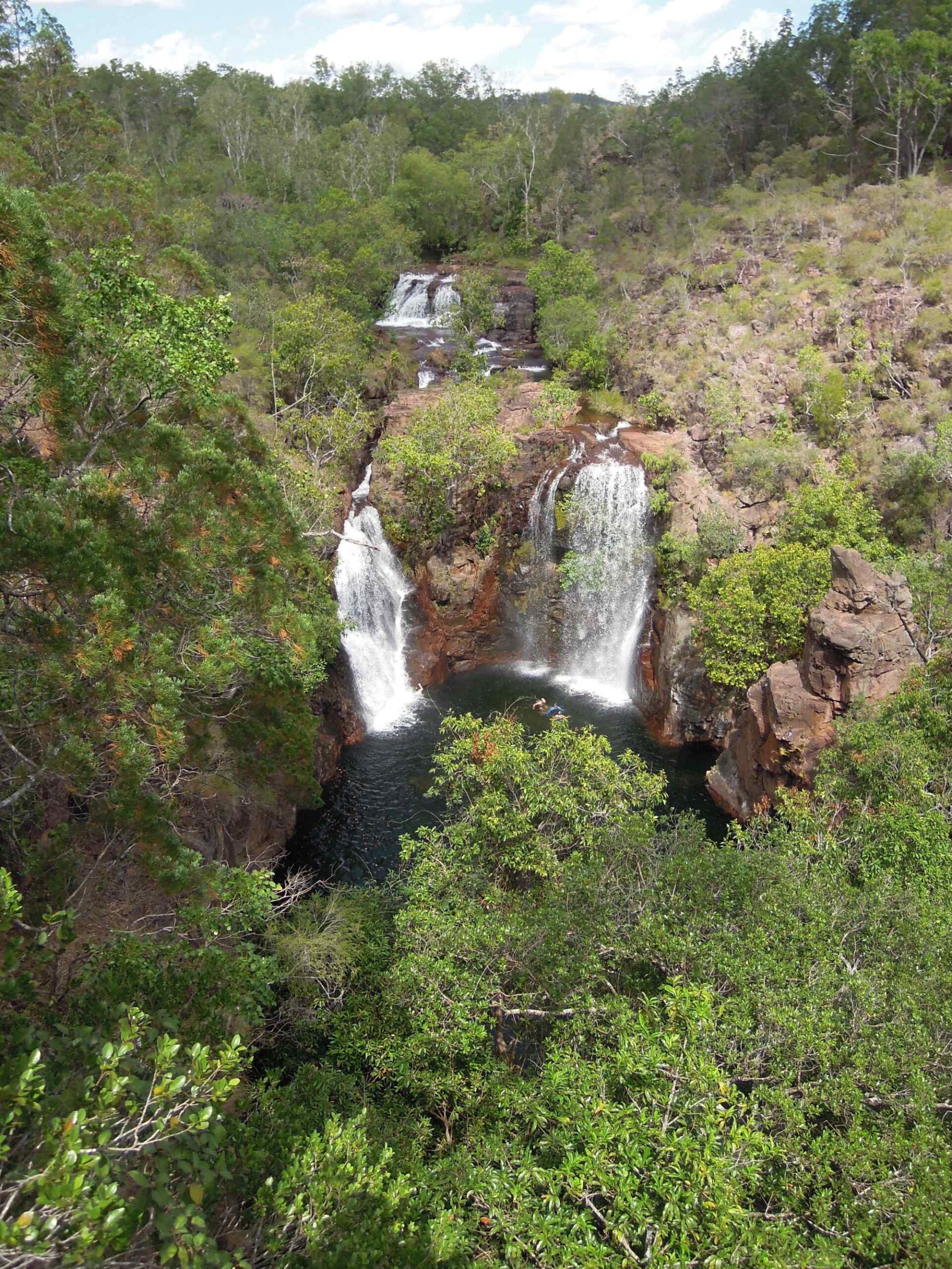 A scenic aerial view of Florence Falls cascading down rocky cliffs into a pristine pool, surrounded by lush eucalyptus forest and native vegetation in Australia's Litchfield National Park. Multiple tiers of waterfalls create a stunning natural landscape in the tropical savanna region.