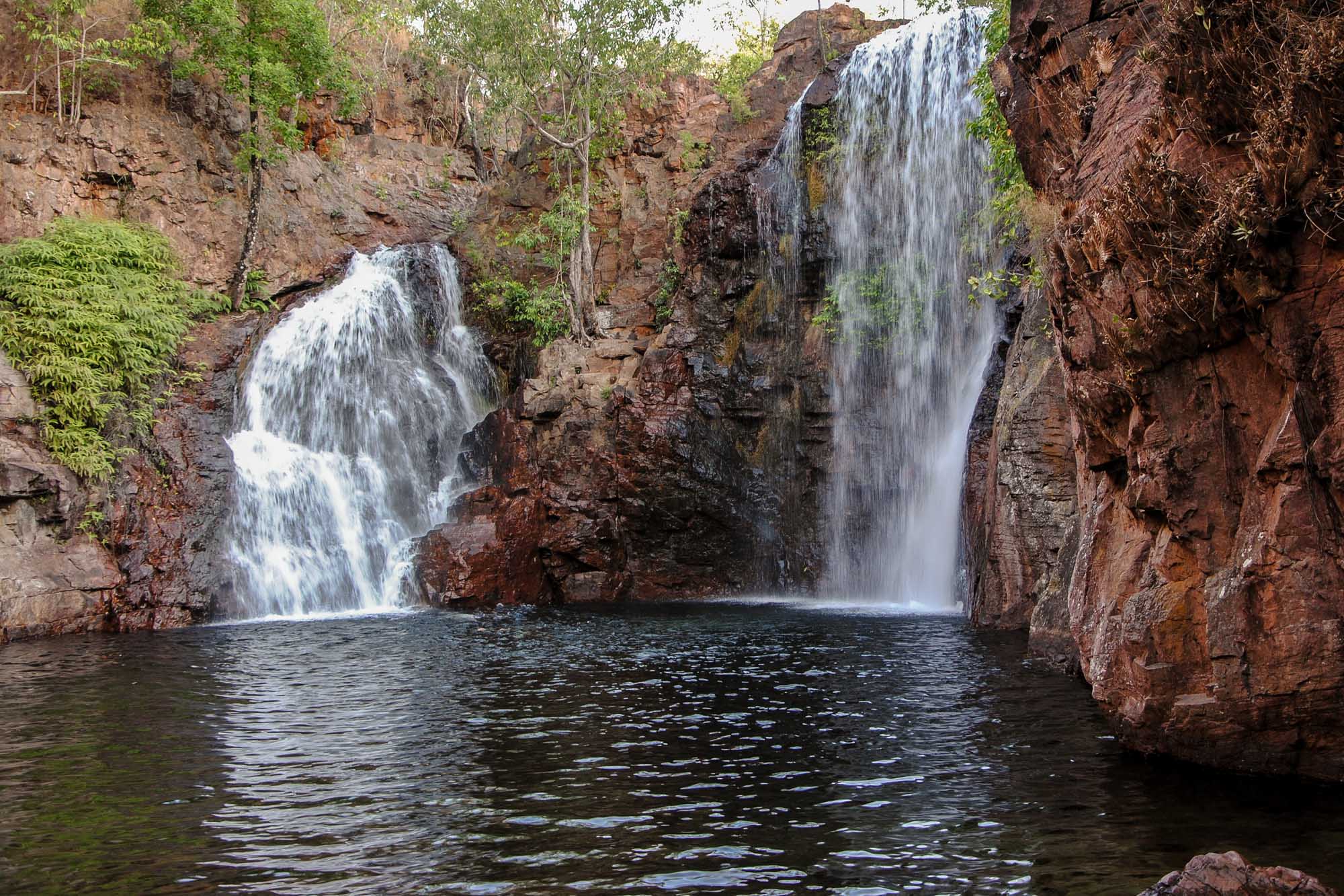 A stunning double waterfall cascades into a pristine plunge pool surrounded by red rock cliffs and lush tropical vegetation in Litchfield National Park. The serene natural pool is framed by towering sandstone formations and verdant bushland, creating an idyllic swimming and photography destination in Australia's Northern Territory.