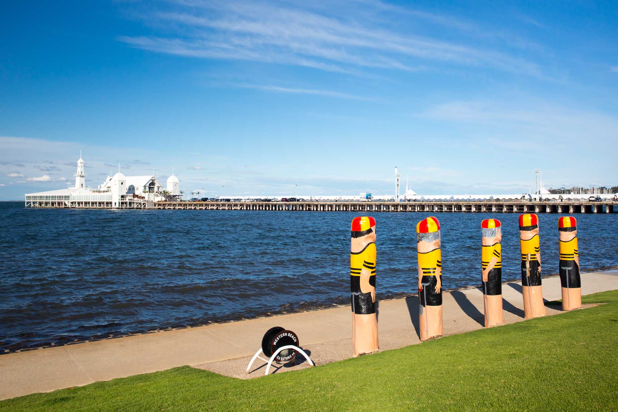 A scenic view of the Geelong waterfront in Victoria, Australia, featuring five distinctive striped bathing boxes painted in yellow and red on the sandy beach, with the historic Geelong pier and its white buildings visible across the blue water under a clear sky.