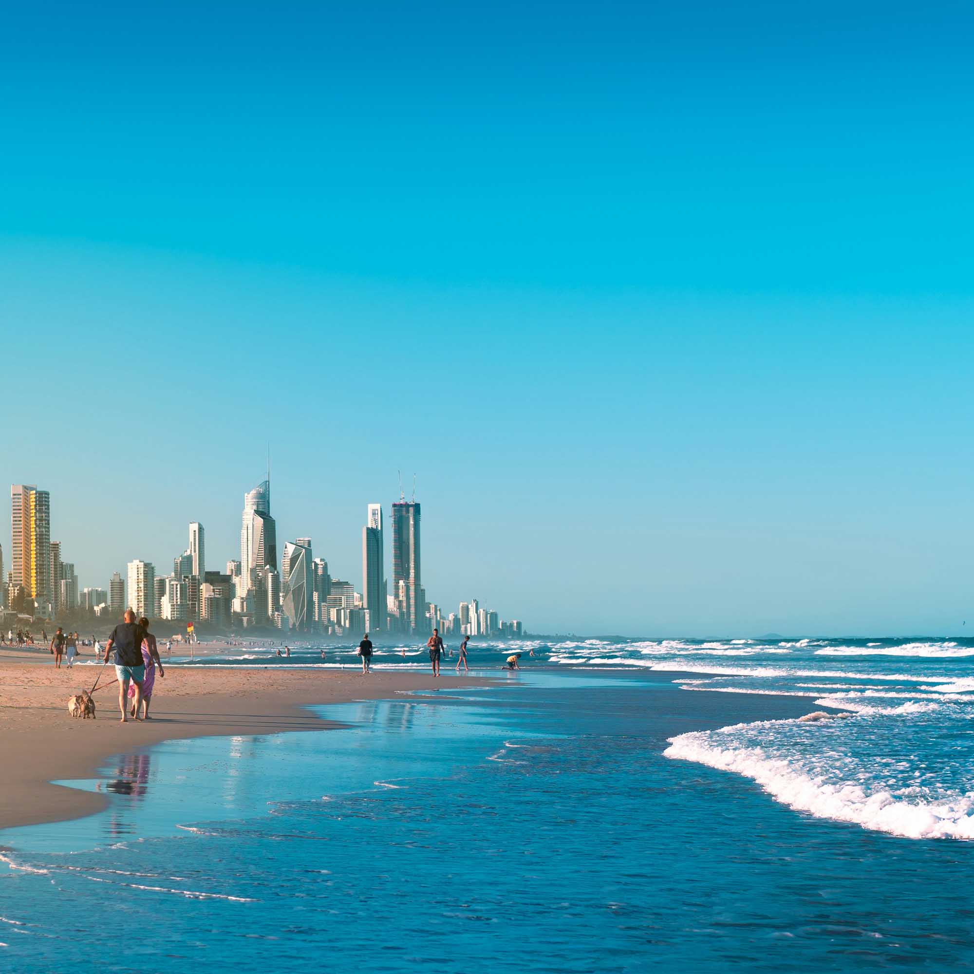 A vibrant coastal scene showcasing the Gold Coast's iconic high-rise skyline against a brilliant turquoise ocean. Beach-goers enjoy the pristine sandy shore and clear blue waters with rolling white-capped waves, under a bright, cloudless sky.