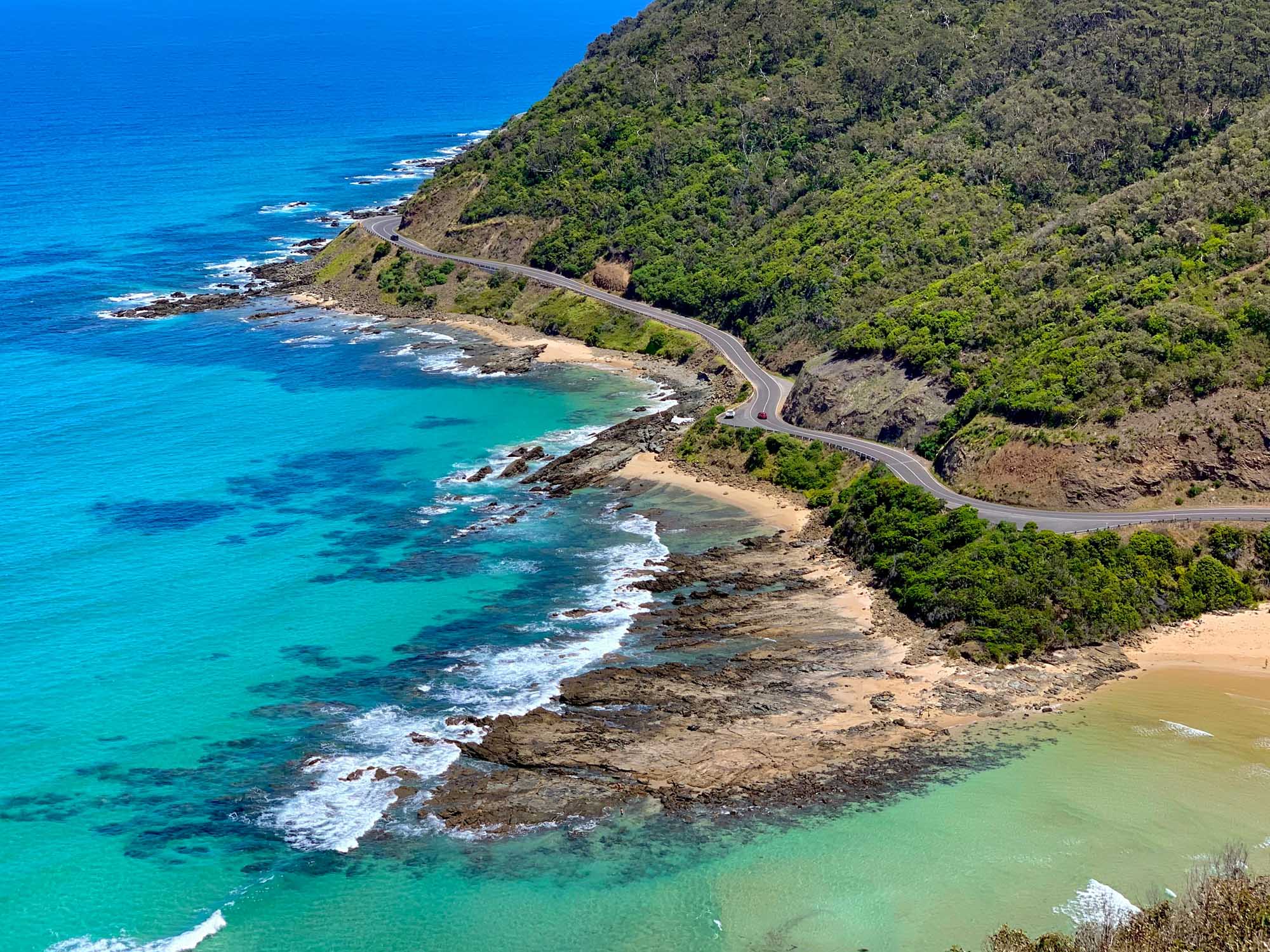 A stunning aerial view of Australia's iconic Great Ocean Road hugging a dramatic coastline with pristine turquoise waters, sandy beaches, and lush green forest-covered cliffs. The winding coastal highway showcases the spectacular meeting of land and sea along Victoria's famous scenic route.