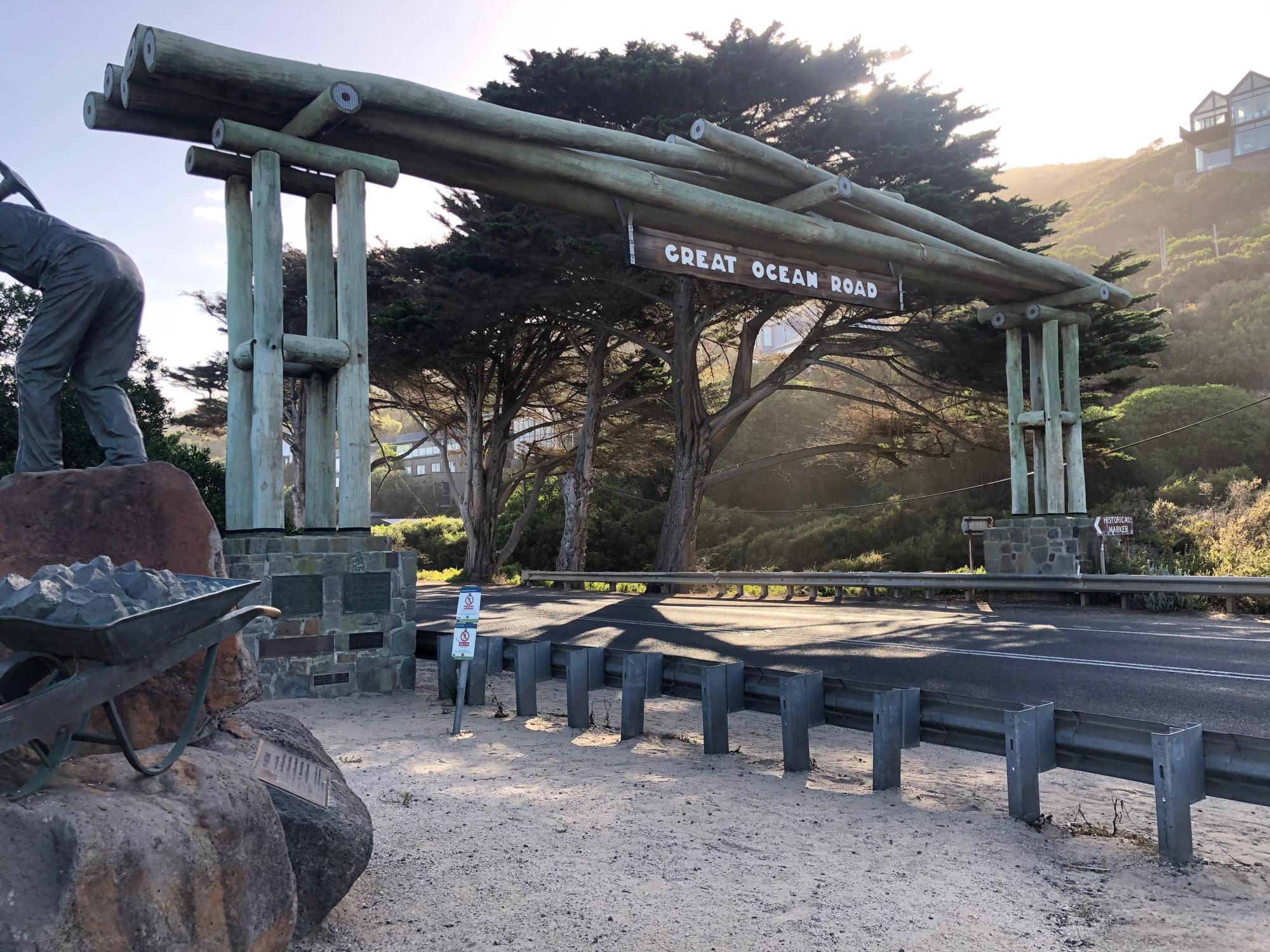 A distinctive rustic gate structure made of weathered green timber with cylindrical support beams frames the entrance to the Great Ocean Road. The gate overlooks a scenic coastal landscape with cypress trees, rolling hills, and a historic building visible on the hillside, captured during golden hour lighting.