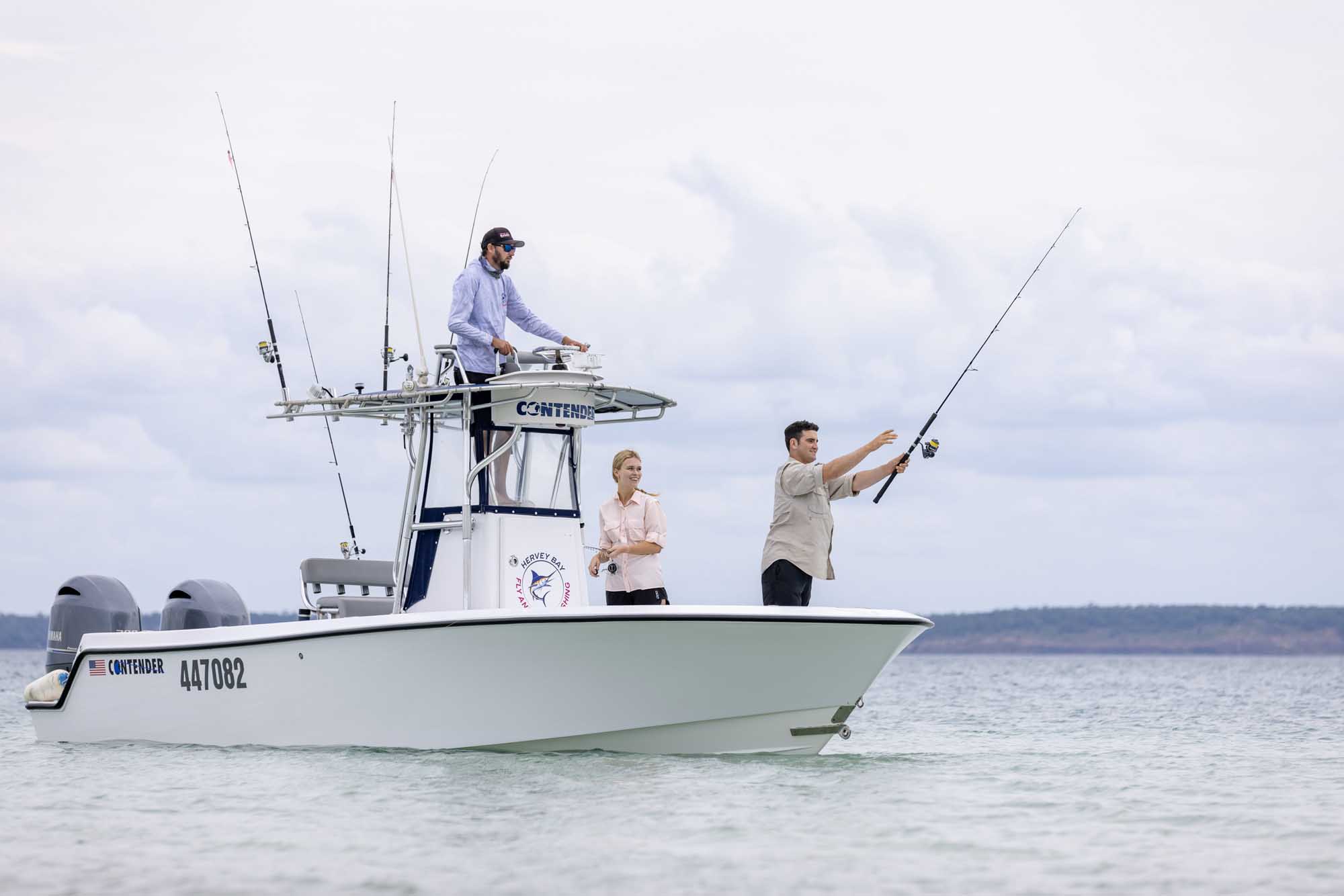 A recreational fishing charter boat named 'Contender' with three anglers aboard in the sheltered waters off K'gari (Fraser Island) near Hervey Bay. One angler actively casts a fishing rod while two others prepare for their turn, with multiple fishing poles rigged on the boat's tower under partly cloudy skies.