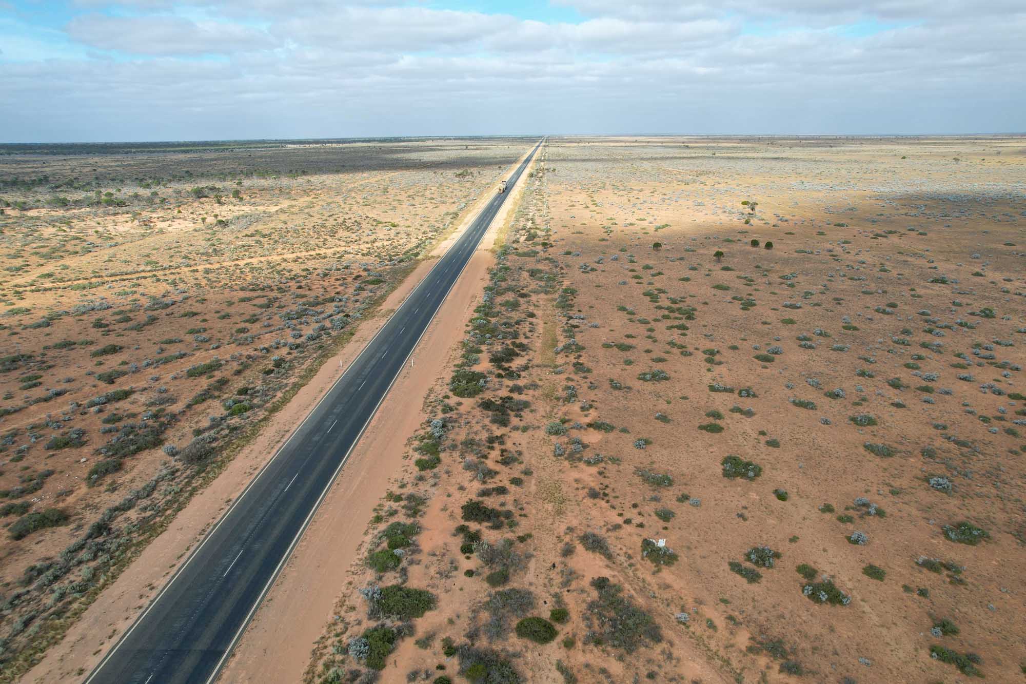 An aerial view of a perfectly straight asphalt highway stretching endlessly across the vast, arid Nullarbor Plain in Australia, with sparse desert vegetation and reddish-brown soil on either side under a partly cloudy sky.