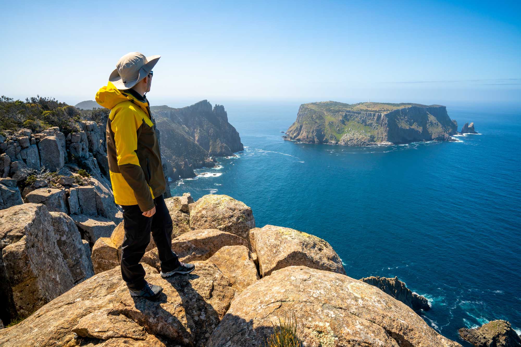 A young hiker in a bright yellow jacket stands on a rocky outcrop overlooking the dramatic coastline of Tasmania's Three Capes Track, with rugged sea cliffs, turquoise waters, and distant rocky islands visible across the pristine blue ocean.