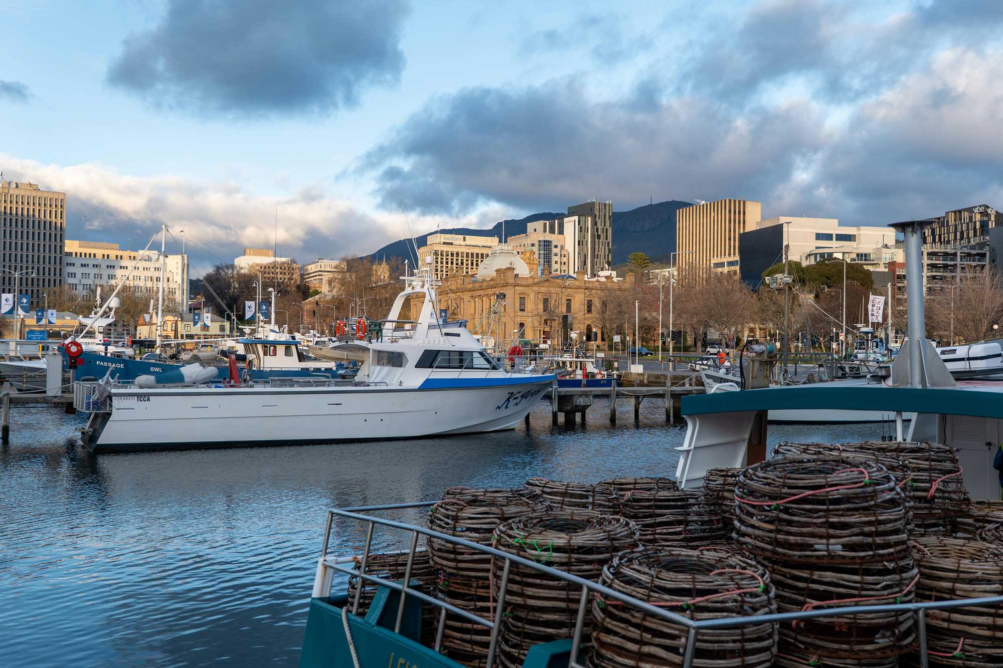 A scenic waterfront view of Hobart's harbor featuring moored fishing vessels with stacked crab pots in the foreground, modern and historic buildings along the waterfront, and mountains visible in the distance under a dramatic cloudy sky. The image captures the working maritime character of Tasmania's capital port.