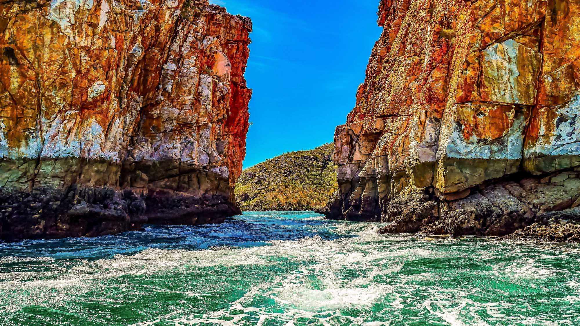 A dramatic coastal gorge featuring towering rust-red and golden rock cliffs framing crystal-clear turquoise waters. The narrow passage between the cliff faces opens to a pristine beach cove, with lush green vegetation visible on the hillside beyond—a signature natural phenomenon of Western Australia's remote Kimberley region.