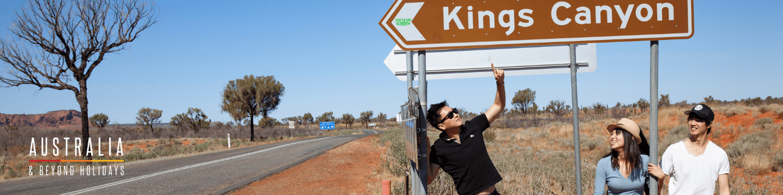 Three tourists posing with a Kings Canyon directional sign along a remote Australian highway, with red earth, sparse vegetation, and clear blue sky characterizing the arid outback landscape. The scene captures the adventurous spirit of road travel through central Australia's iconic natural attractions.