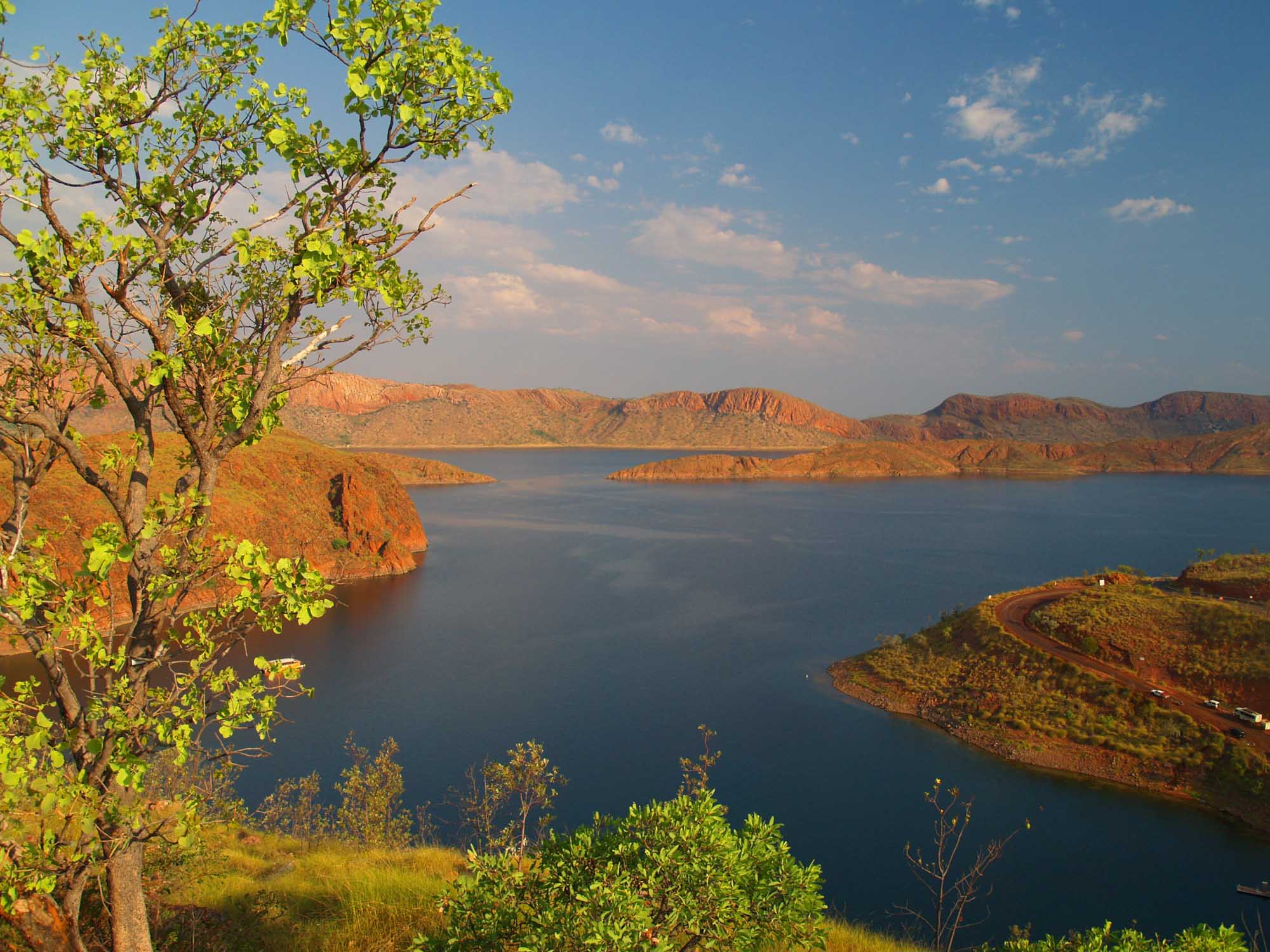A stunning golden hour view of Lake Argyle in the Kimberley region of Western Australia, showcasing rugged red rock formations and deep blue water under a partly cloudy sky. Native vegetation frames the foreground, highlighting the natural beauty of this remote inland water body.