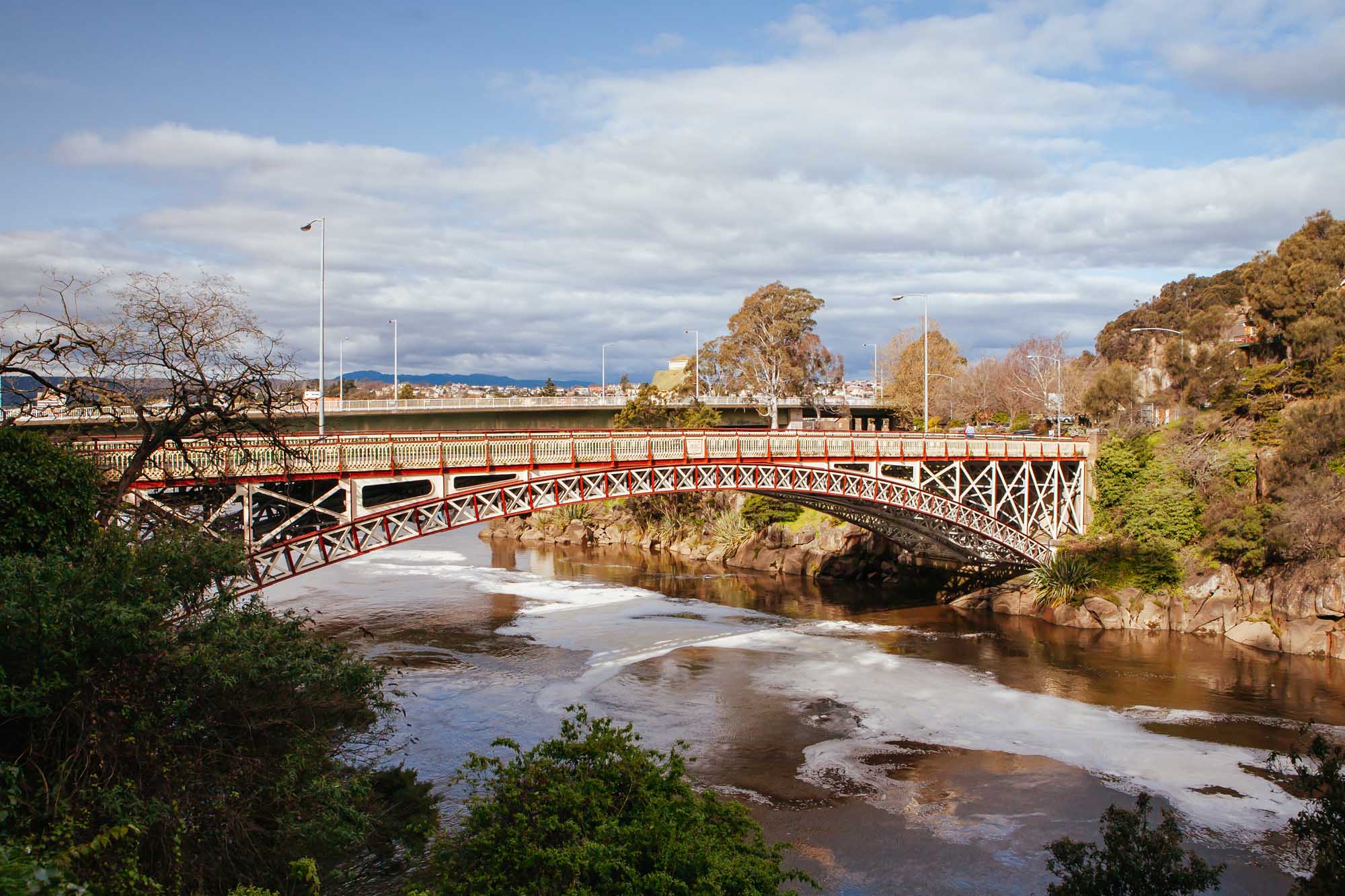 A striking red and white Victorian-era pedestrian bridge spans the Cataract Gorge at First Basin in Launceston, Tasmania. The ornate iron lattice bridge arches gracefully over the tidal river with its exposed mudflats and rocky outcrops, set against a backdrop of native Australian bushland and mountains under a partly cloudy sky.