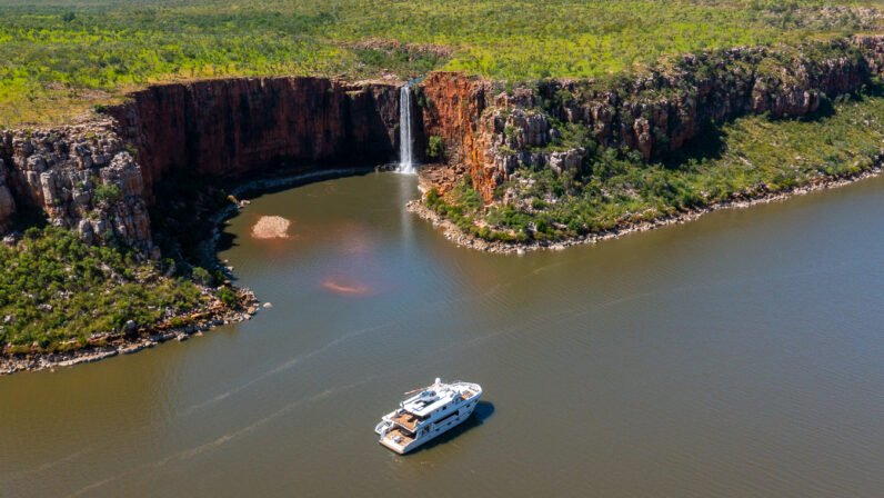 An aerial view of the Kimberley Quest small expedition vessel anchored in a pristine river gorge, with a spectacular waterfall cascading down red rock cliffs and lush tropical vegetation covering the surrounding landscape in Western Australia's Kimberley region.