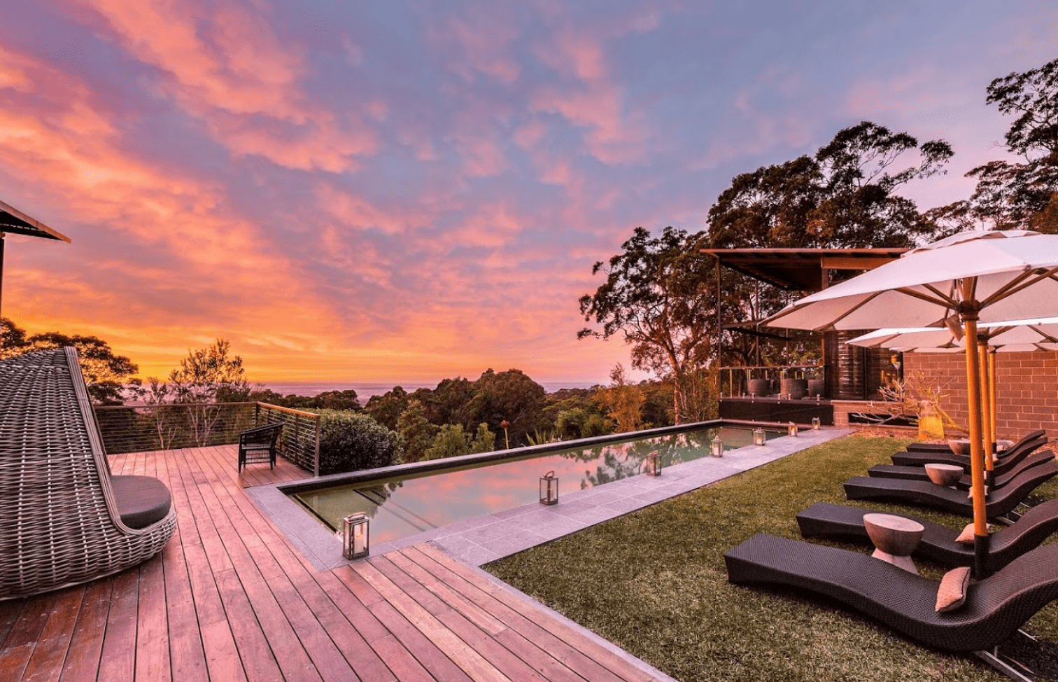 An expansive luxury resort property featuring an elegant infinity pool overlooking a sprawling landscape, with manicured lawns, modern lounge chairs with umbrellas, and contemporary architecture bathed in the warm glow of a golden sunset sky. The scene captures the epitome of high-end hospitality and relaxation with panoramic vistas across forested hills and valleys.