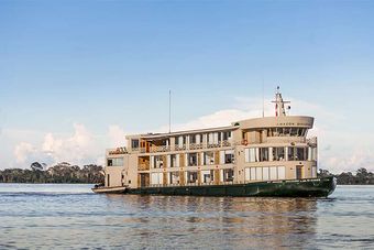 An elegant multi-story riverboat with classical colonial architecture and ornate white railings floats on calm blue water under clear skies. The vessel features distinctive mansion-like design with multiple decks, large windows, and a tall antenna, suggesting it serves as a luxury floating accommodation or cruise vessel on a major river system.