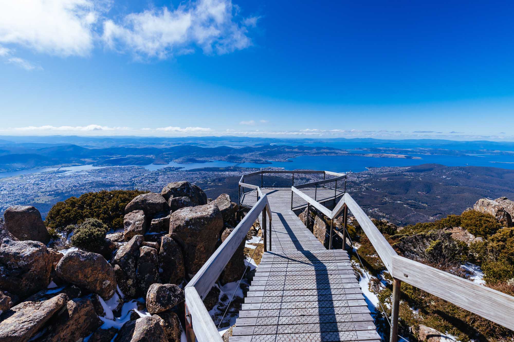A scenic wooden boardwalk observation platform at the summit of Mt Wellington with panoramic views over Hobart, Tasmania, featuring the Derwent River, surrounding lakes, and distant mountains under a clear blue sky on a crisp spring morning.