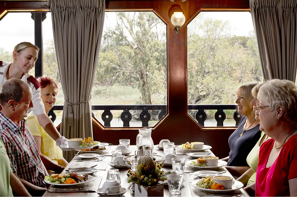 An elegant formal dining room aboard the Murray Princess river cruise ship, featuring guests enjoying a meal at a long table with fine place settings, white tablecloths, and large windows overlooking lush green landscapes and eucalyptus trees along the Murray River.