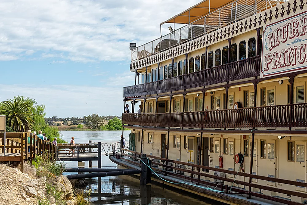 The Murray Princess, a multi-deck riverboat, is docked along the Murray River in South Australia. The distinctive yellow and cream-colored paddlewheel vessel features multiple levels with ornate railings and wide viewing decks, characteristic of classic Australian river cruising vessels.