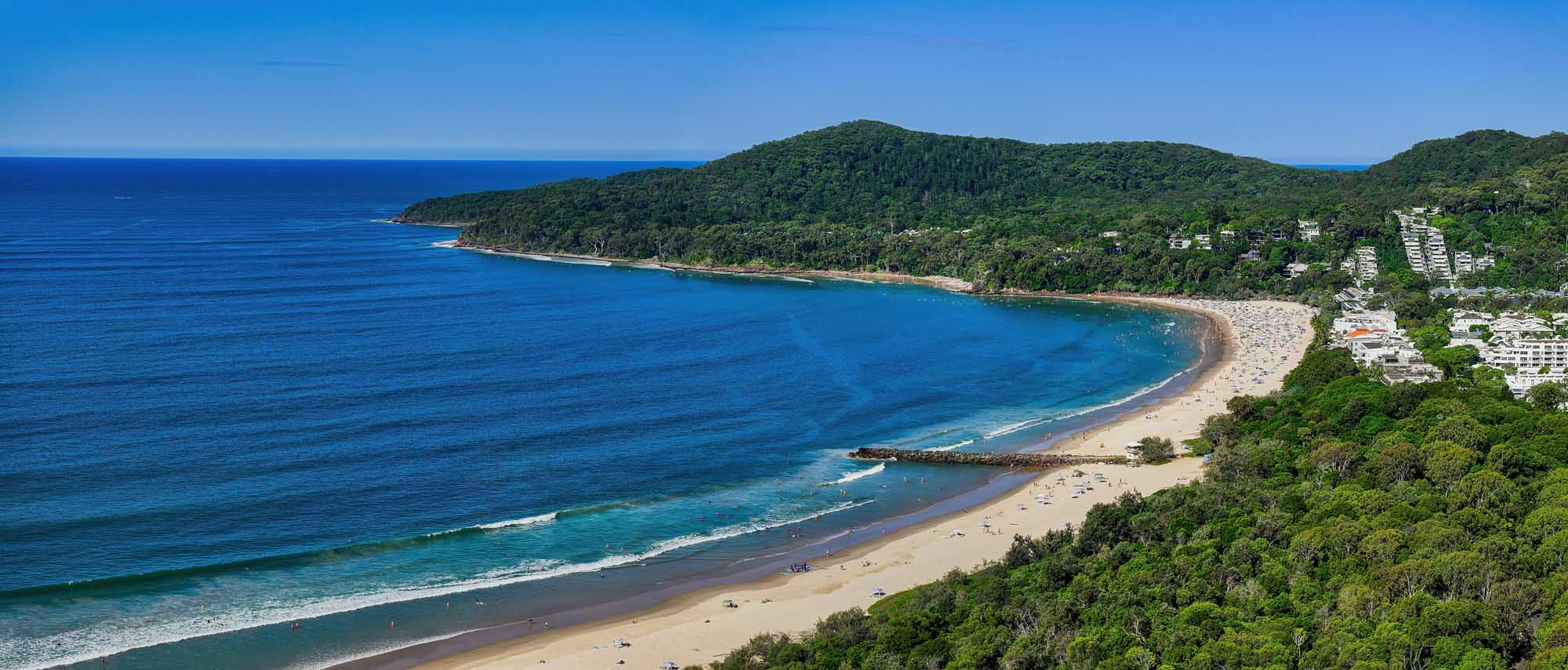 An aerial panoramic view of Noosa Heads main beach in Queensland, Australia, featuring a pristine sandy shoreline framed by turquoise waters, with Noosa National Park's densely forested headland in the background and beachfront residential development visible on the right.