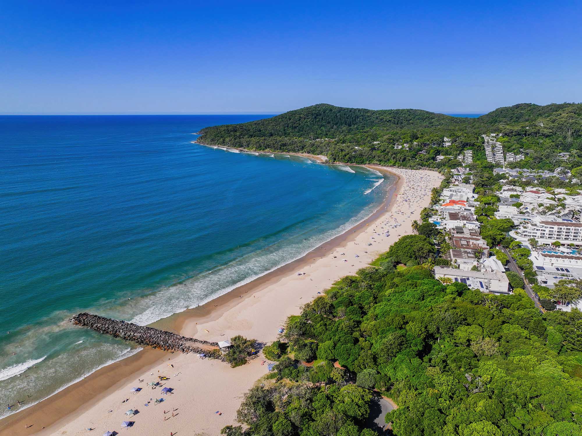 An aerial photograph of Noosa Heads main beach in Queensland, Australia, showing pristine golden sand, crystal-clear turquoise waters, and lush green bushland of Noosa National Park. The image captures the beach's crescent shape with residential development nestled among dense coastal vegetation, creating a striking contrast between natural landscape and modern settlement.