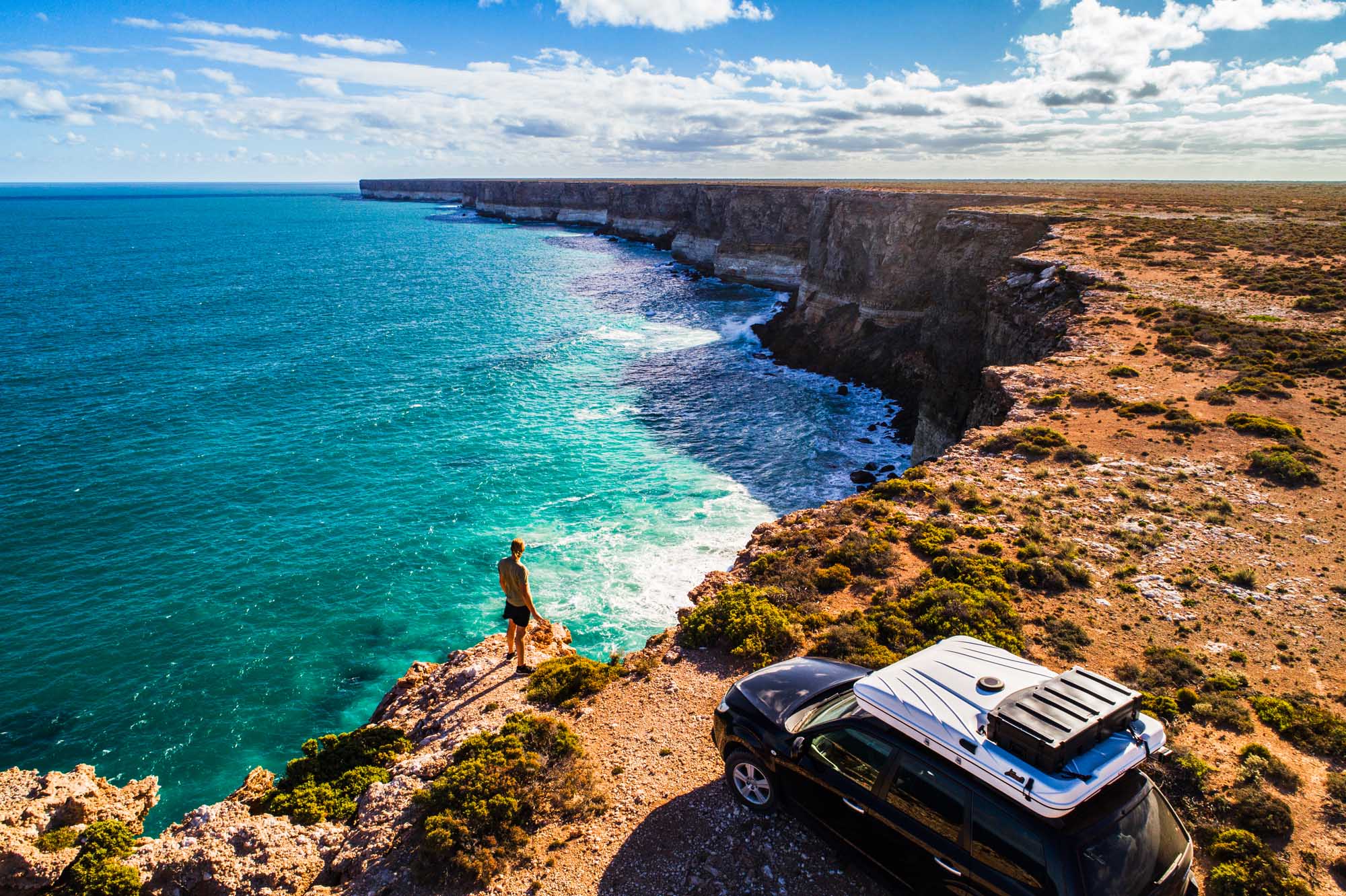 An aerial view of a black SUV with a rooftop camper parked on rugged coastal cliffs overlooking the turquoise waters of the Great Australian Bight. A lone traveler stands at the cliff's edge, gazing across the dramatic limestone formations and pristine seascape under a partly cloudy sky.