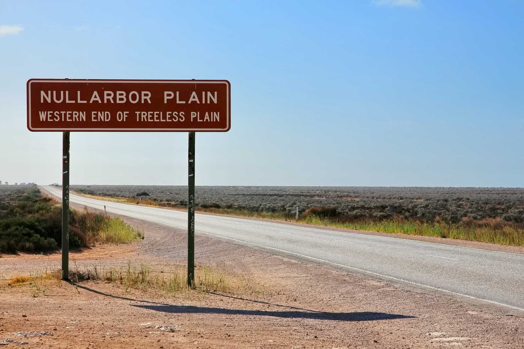 A brown directional sign marking the western end of the treeless Nullarbor Plain stands beside a straight highway stretching across an arid, sparse landscape under clear blue skies. The vast, flat terrain with scattered low vegetation exemplifies the remote and desolate character of this Australian outback region.