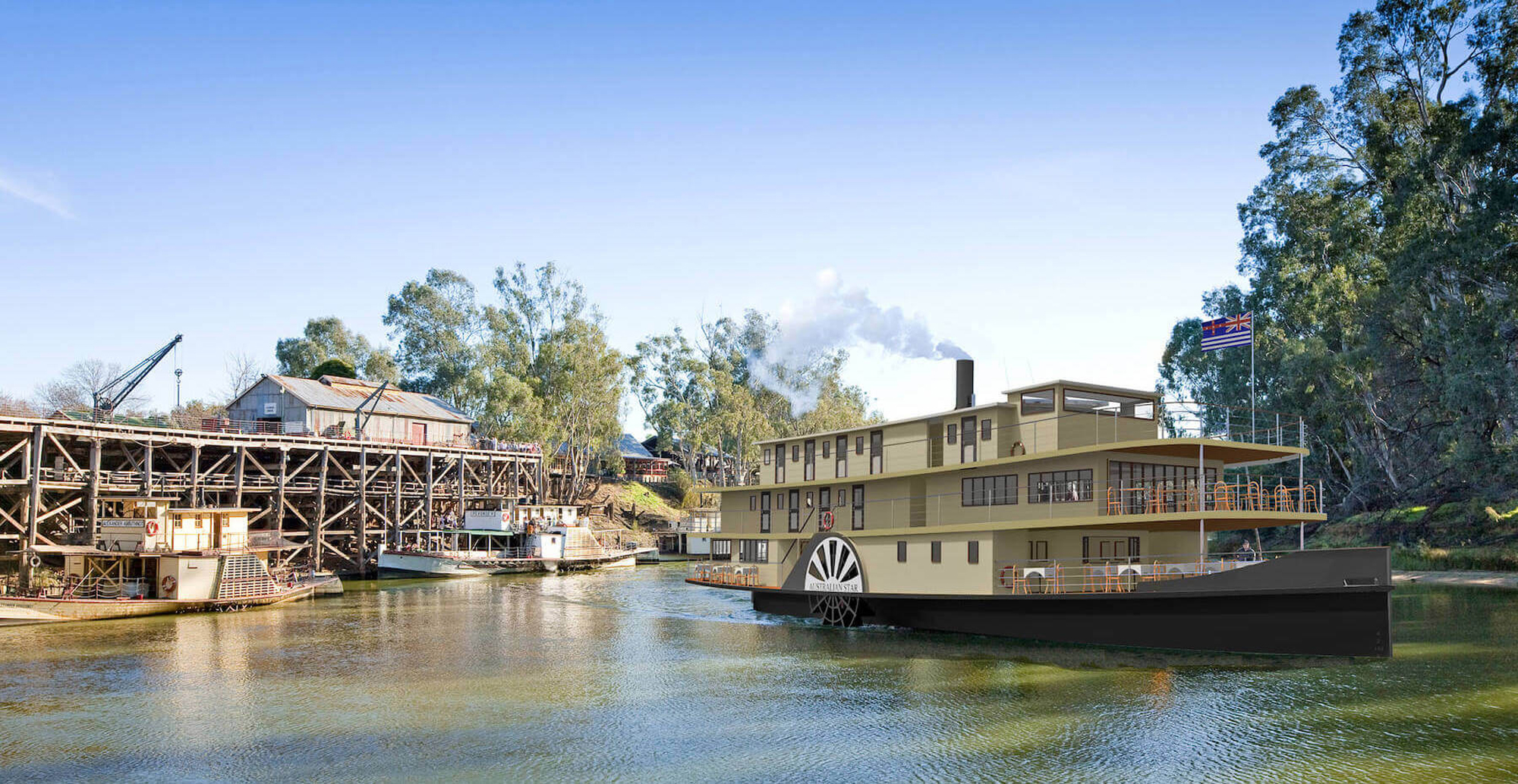 A classic multi-deck paddlewheel riverboat with cream and dark hull coloring is moored at a historic riverside wharf featuring vintage timber infrastructure, loading equipment, and period buildings. The scene captures the heritage of river cruise tourism on what appears to be the Murray River in Australia.