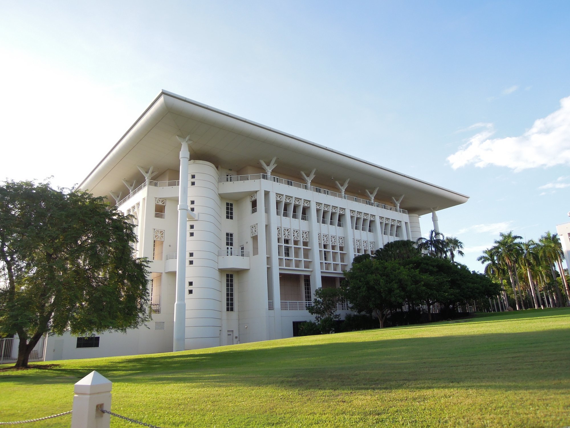 A striking white modernist government building featuring distinctive curved architectural elements, broad overhanging eaves, and decorative screen panels. The structure is set on manicured grounds with palm trees and tropical landscaping, exemplifying mid-20th century Australian institutional design.