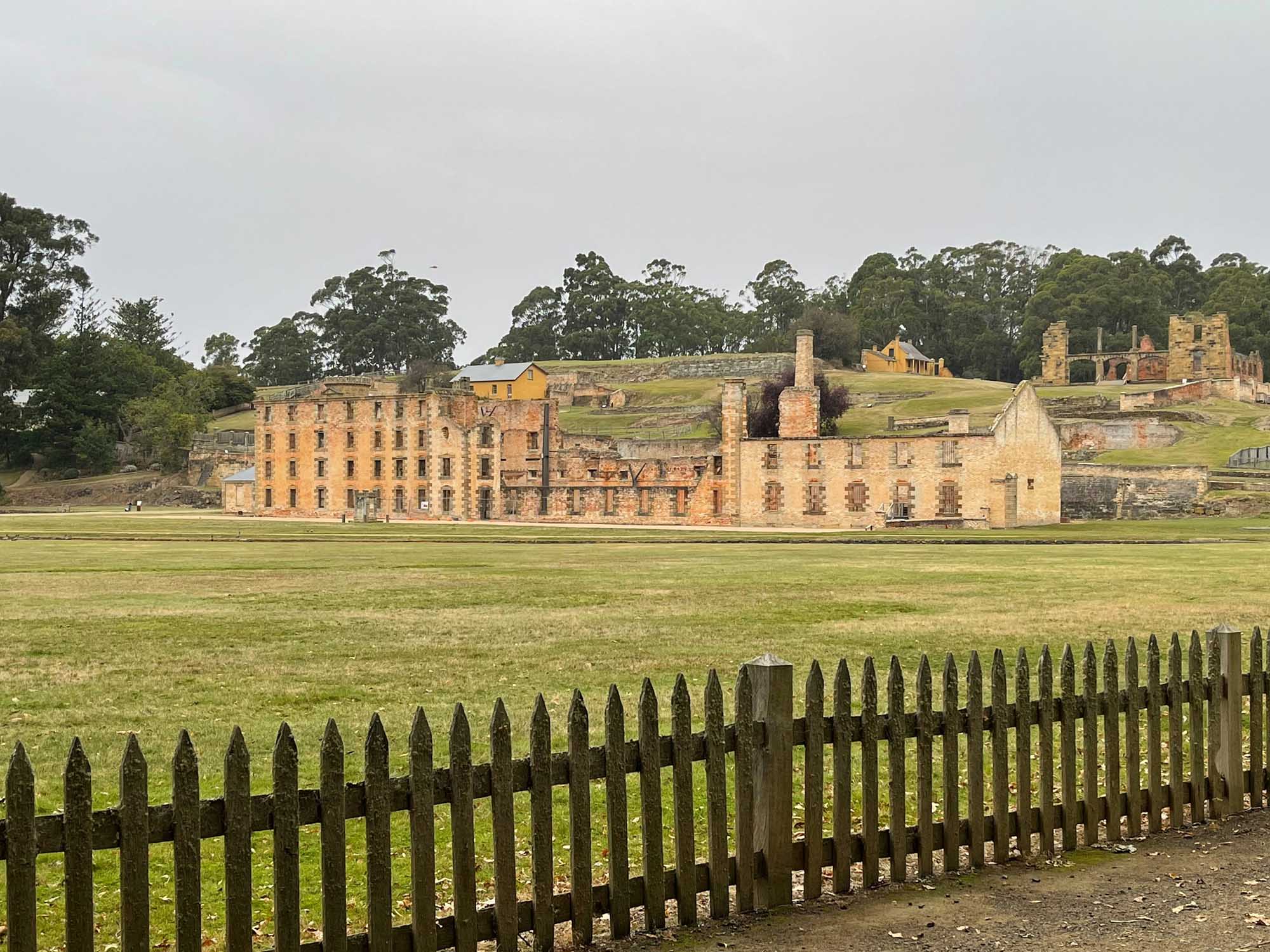 A panoramic view of the historic Port Arthur penal settlement in Tasmania, Australia, featuring well-preserved sandstone buildings and ruins scattered across a grassy landscape, with native forest in the background. The wooden fence in the foreground frames the 19th-century convict prison complex, a UNESCO World Heritage site.
